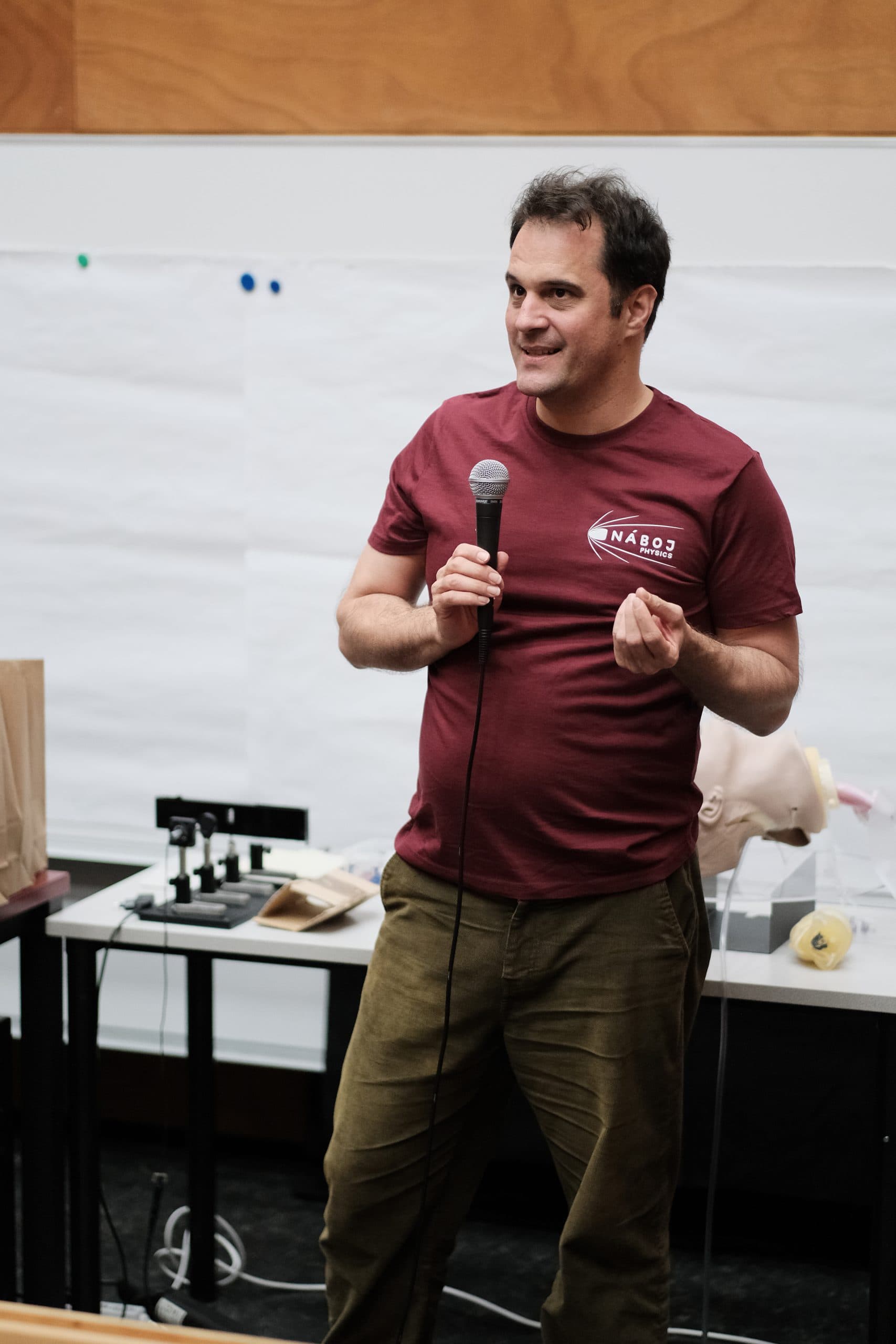 Ein Mann in einem kastanienbraunen T-Shirt hält ein Mikrofon in der Hand und spricht vor einer Tafel und einem Tisch mit verschiedenen Objekten, darunter ein Schaufensterpuppenkopf, in einem Klassenzimmer oder einer Präsentationsumgebung.