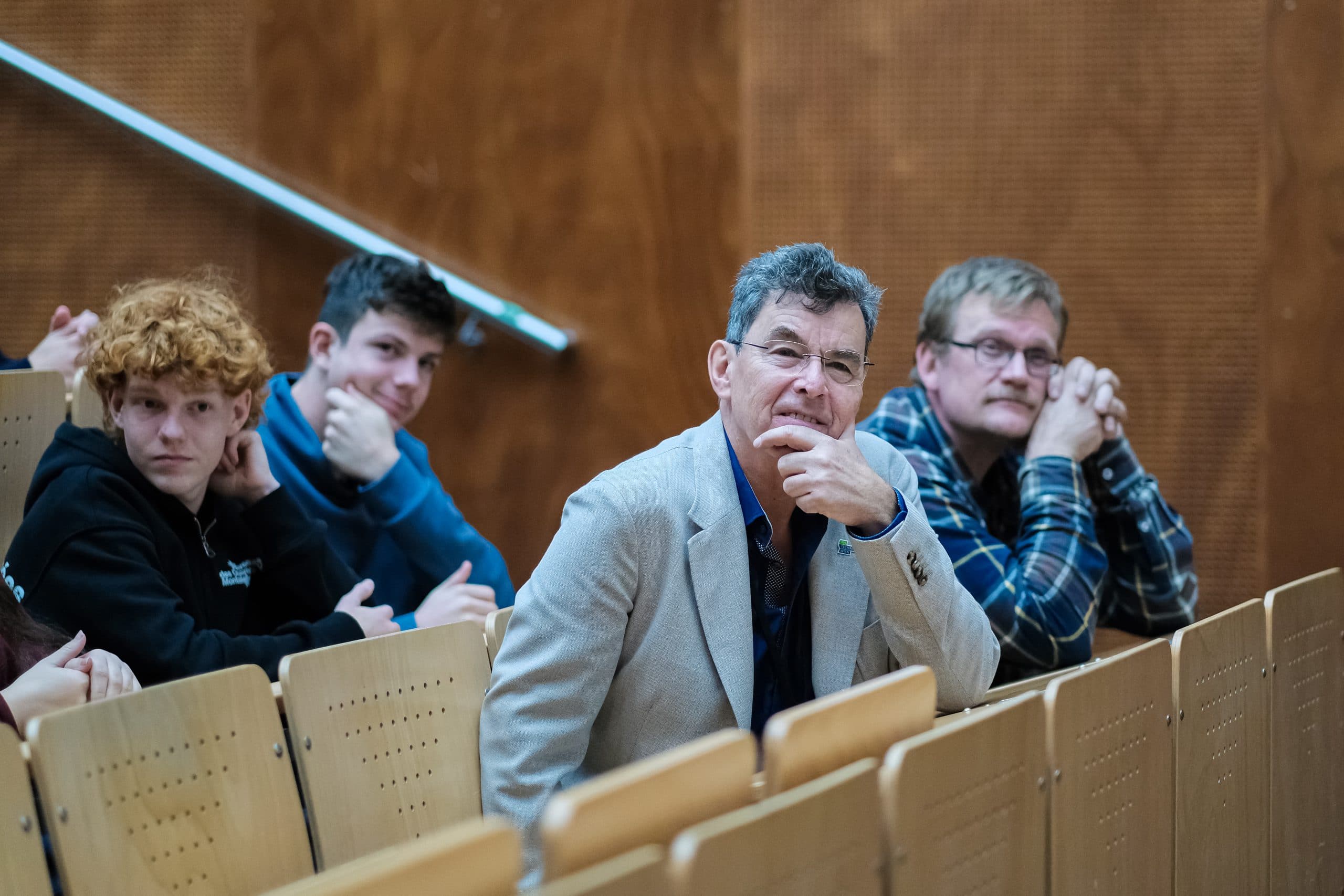 Eine Gruppe von Männern sitzt auf hölzernen Sitzen im Auditorium, stützt das Kinn auf die Hände und blickt aufmerksam nach vorne. Der Hintergrund ist eine braune, strukturierte Wand.
