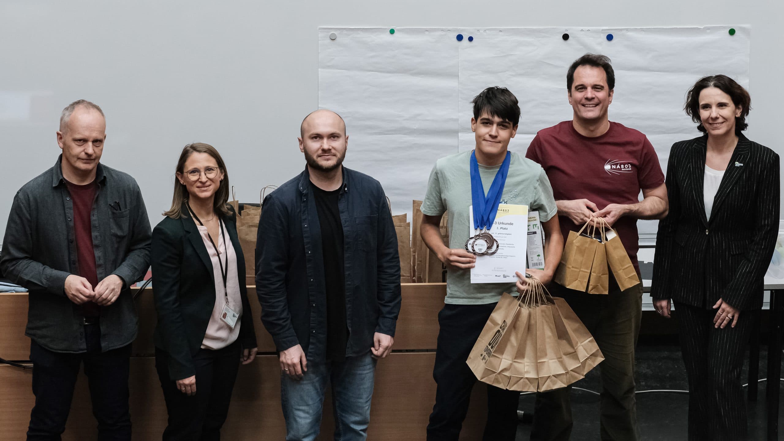 Ein junger Mann in der Mitte hält eine Urkunde, Medaillen und braune Papiertüten in der Hand, während die anderen lächelnd an den Seiten stehen. Hinter ihnen sind leere Blätter an die Wand gepinnt.