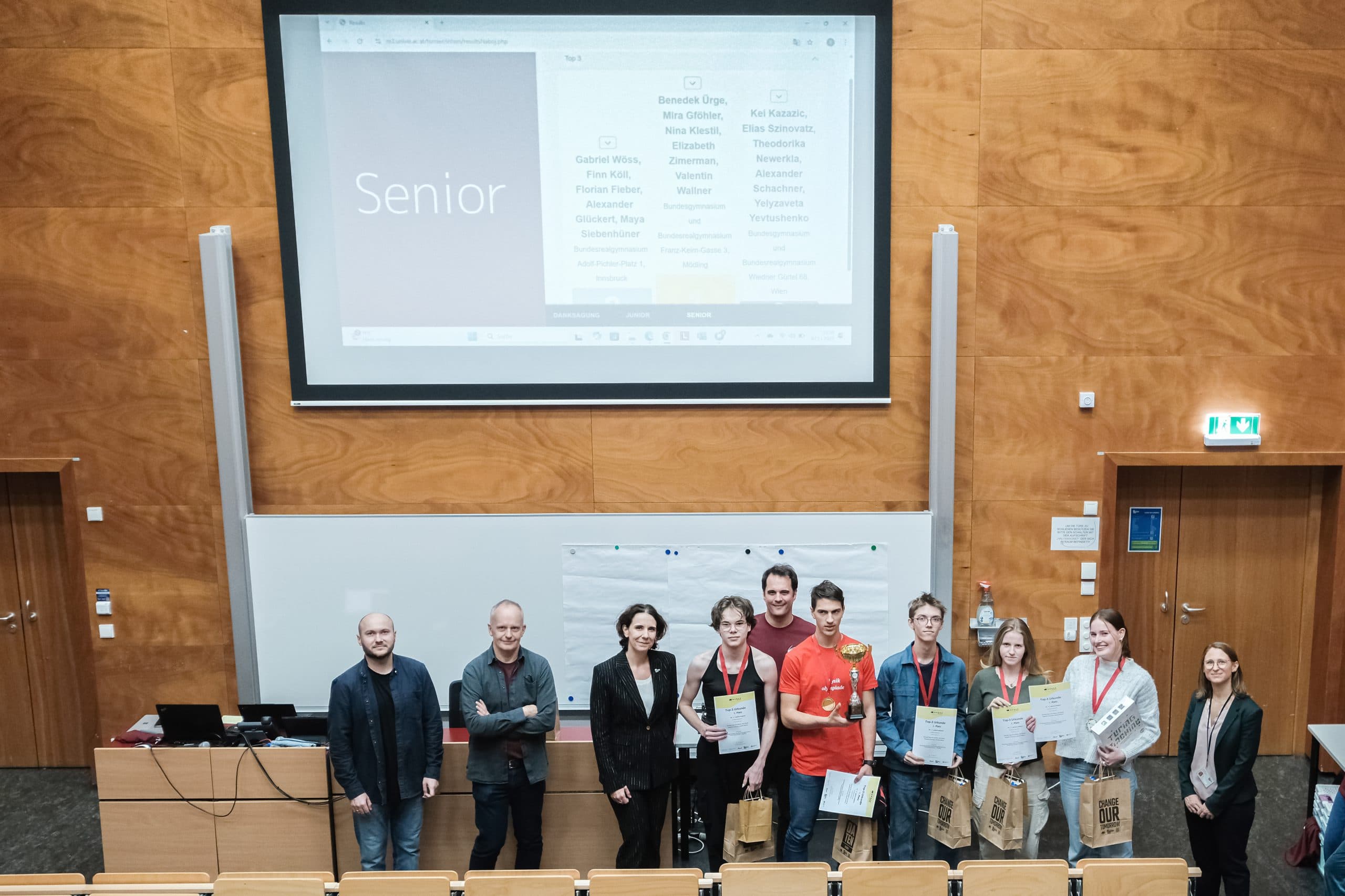 Eine Gruppe von Personen steht am Eingang eines Hörsaals und hält Urkunden und Geschenktüten in der Hand, einige von ihnen tragen Medaillen. Auf einem Bildschirm hinter ihnen erscheinen das Wort Senior und eine Liste von Namen.
