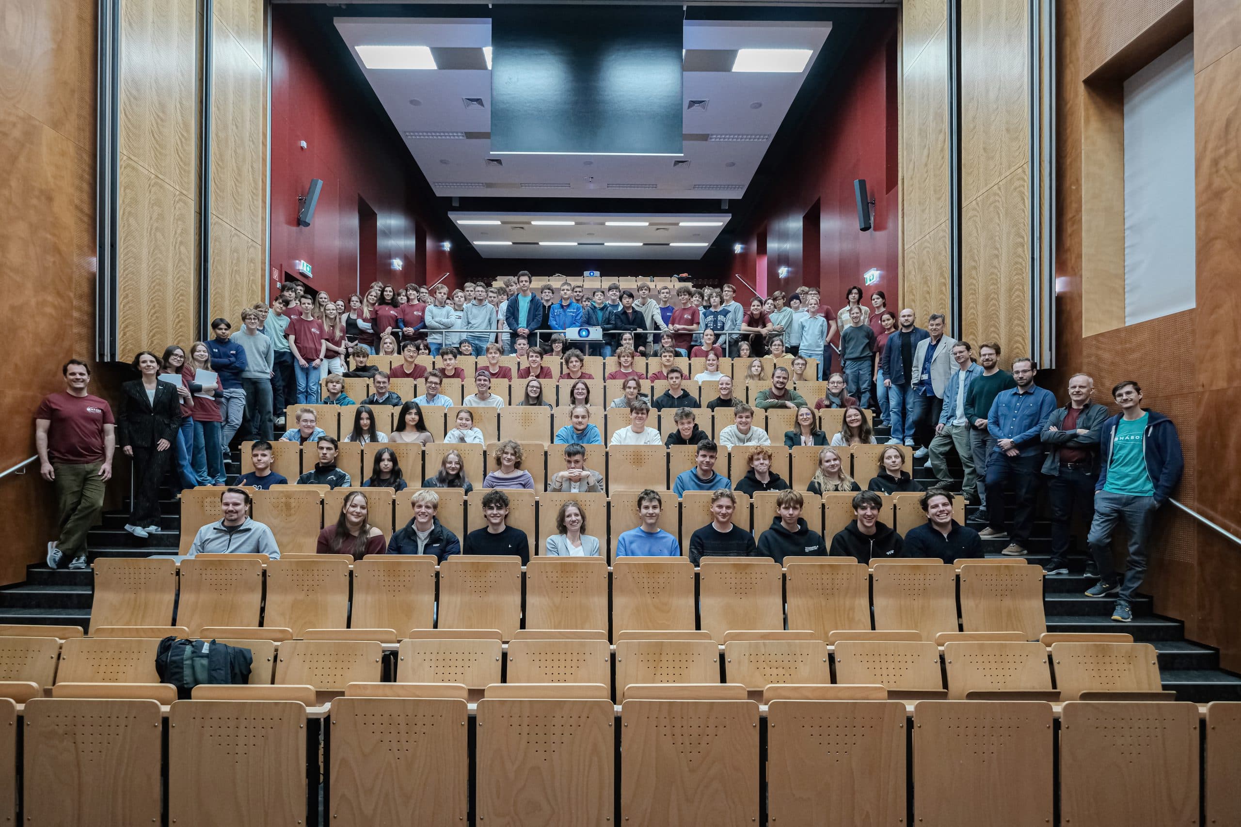Eine große Gruppe von Menschen steht und sitzt in einem Auditorium mit Reihen leerer Holzsitze im vorderen Bereich und stellt sich für ein Gruppenfoto auf. Der Raum hat rote Wände und eine hohe, beleuchtete Decke.