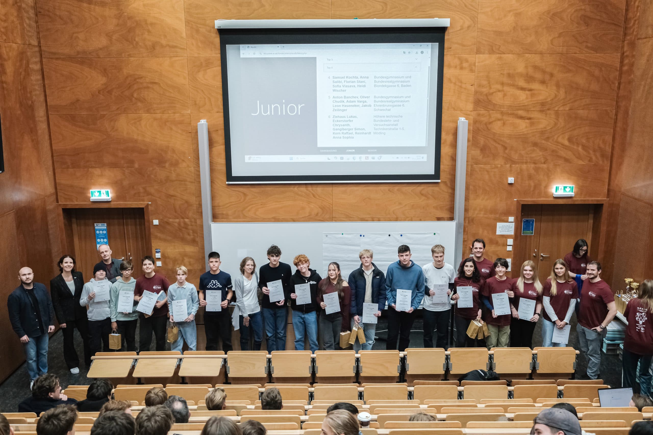 Eine Gruppe Jugendlicher und Erwachsener steht mit Urkunden und Geschenktüten in der Hand vor einem Hörsaal, hinter ihnen ist auf einer großen Leinwand das Wort 'Junior' zu sehen. Die Schüler sitzen den Gewinnern gegenüber.