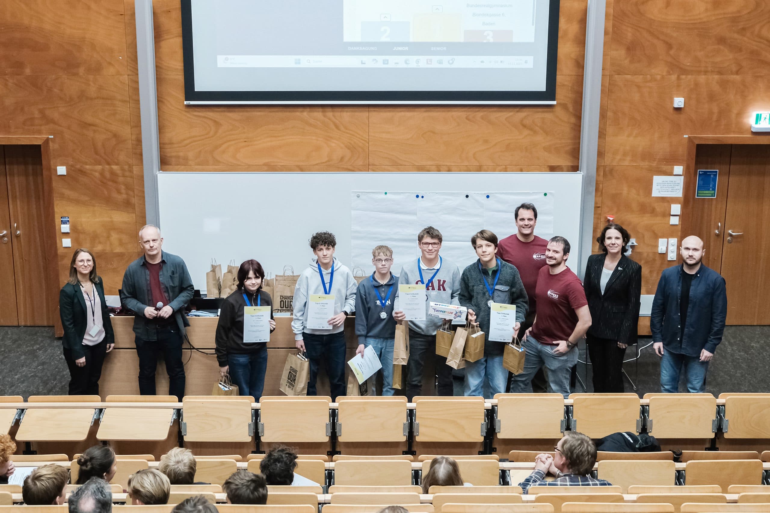 Eine Gruppe von Personen, darunter Studenten mit Zeugnissen und Medaillen, steht am Eingang eines Hörsaals. Erwachsene stehen auf beiden Seiten, und die Zuhörer sitzen ihnen gegenüber. Hinter ihnen sind eine Projektionsfläche und eine Tafel zu sehen.