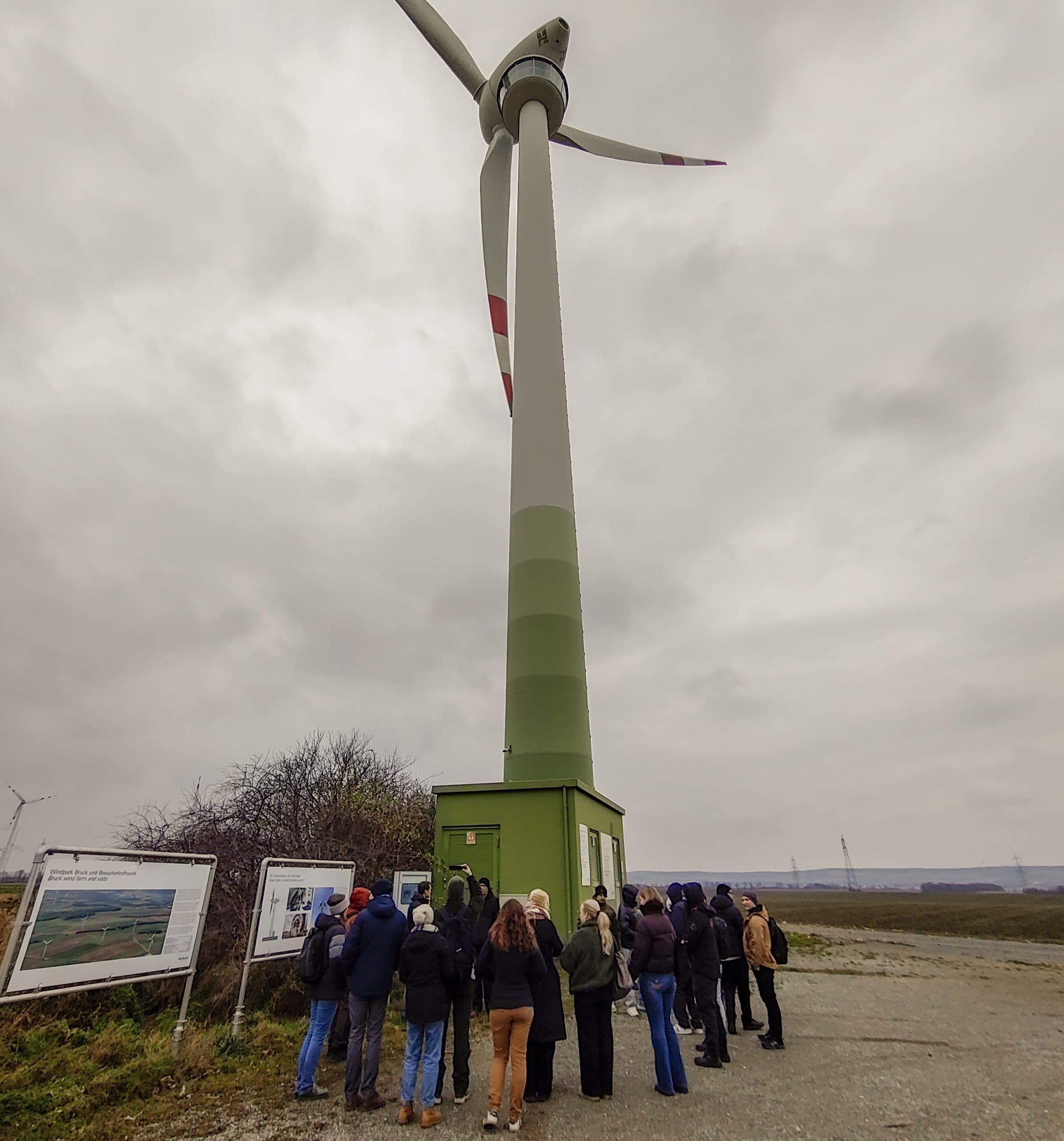 Eine Gruppe von Menschen steht an einem bewölkten Tag in der Nähe des Sockels einer hohen Windkraftanlage und betrachtet die Informationstafeln neben der Anlage in einer offenen Landschaft.