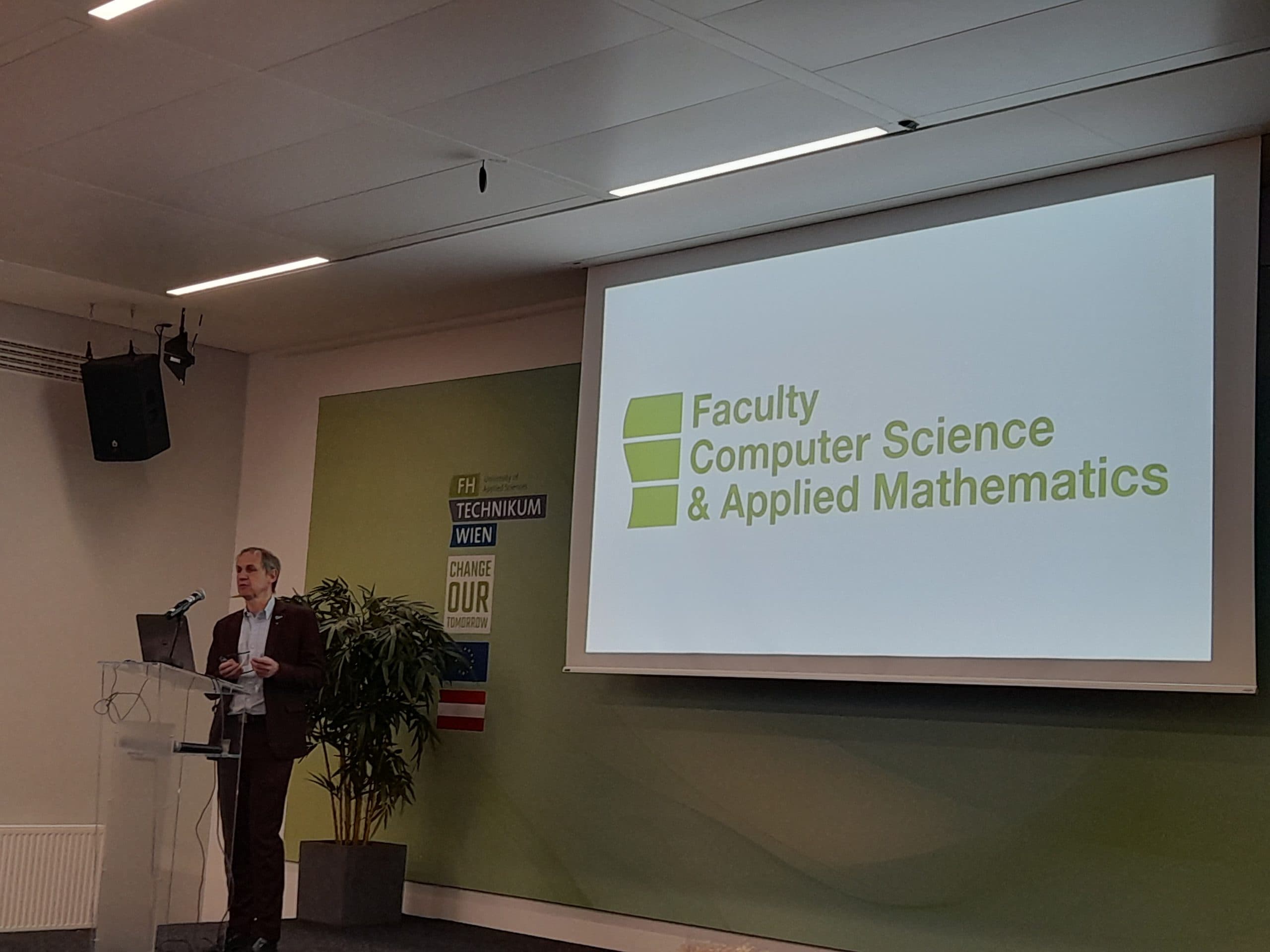 Ein Mann im Anzug steht an einem Podium und hält einen Vortrag. Hinter ihm zeigt eine Projektionsfläche das Logo und den Namen 'Fakultät für Informatik und angewandte Mathematik'. Die Umgebung scheint ein moderner Hörsaal zu sein.