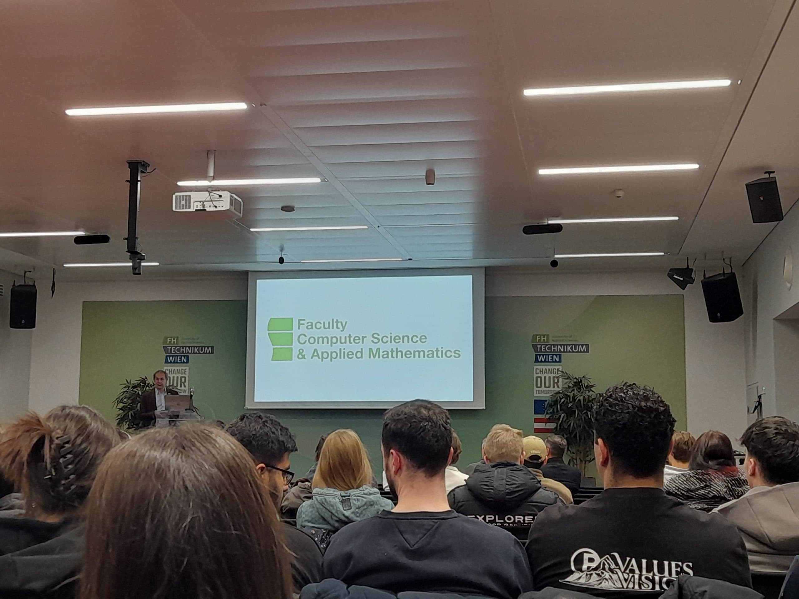 Die Zuhörer sitzen in einem Hörsaal vor einem Bildschirm, auf dem die Fakultät für Informatik und angewandte Mathematik abgebildet ist, mit Bannern an den Wänden und Deckenleuchten über dem Kopf.