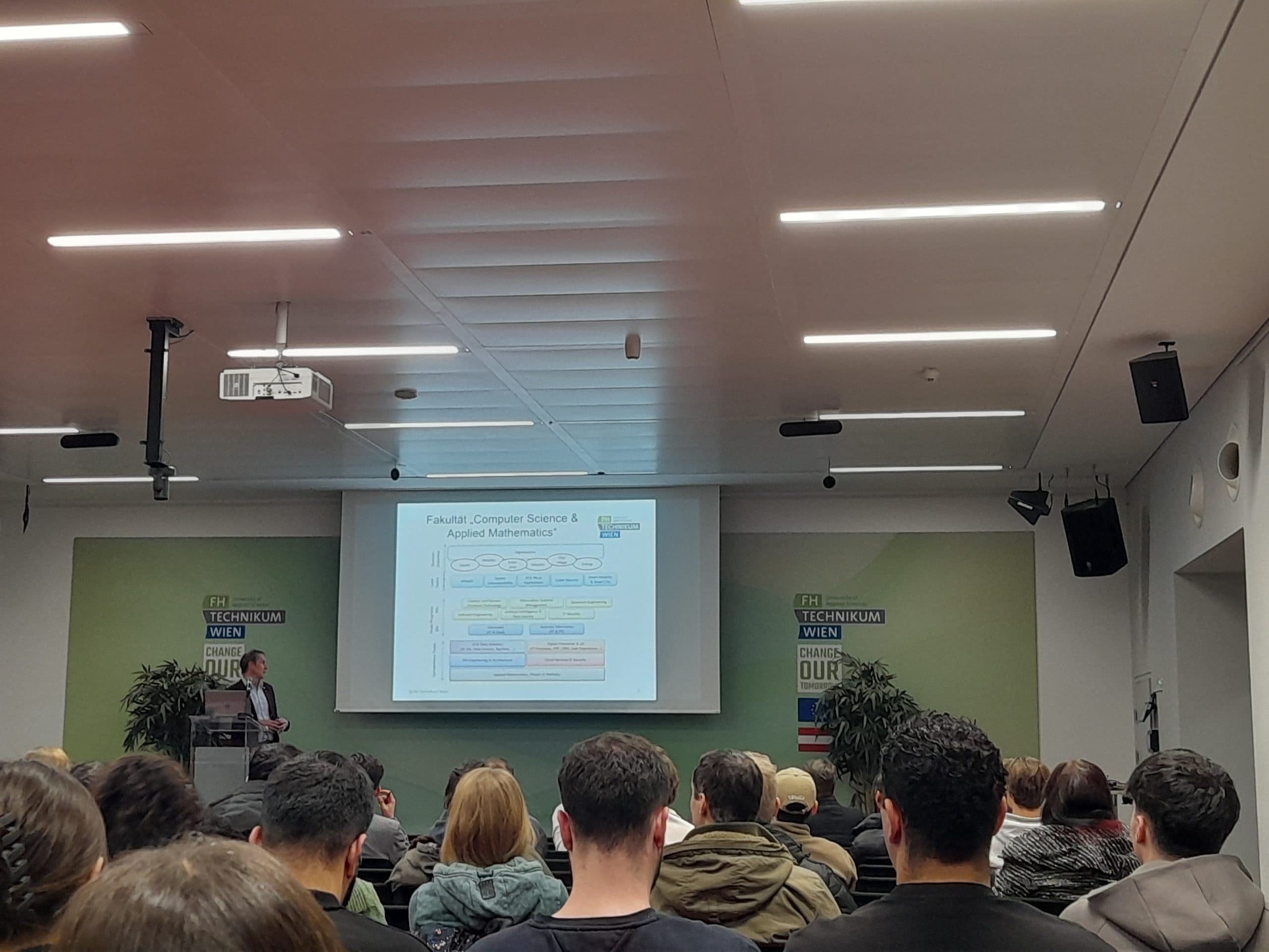 Ein Dozent steht an einem Podium und präsentiert dem Publikum in einem modernen Klassenzimmer eine Folie mit dem Titel Fakultät für Informatik und angewandte Mathematik. Die Folie zeigt ein Diagramm und farbige Kästchen. Die Zuhörer blicken aufmerksam nach vorne.