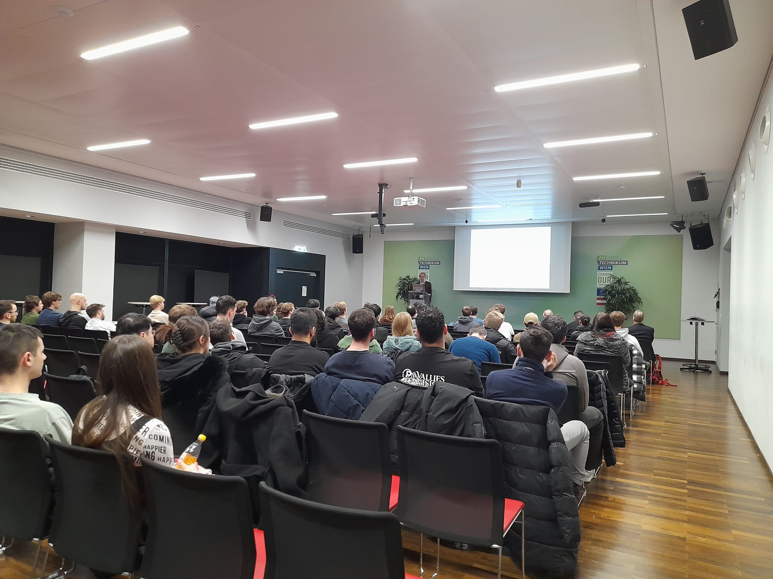 In einem modernen, gut beleuchteten Konferenzraum mit Holzfußboden sitzt eine Gruppe von Personen in Stuhlreihen vor einem Lautsprecher und einer Projektionsfläche.