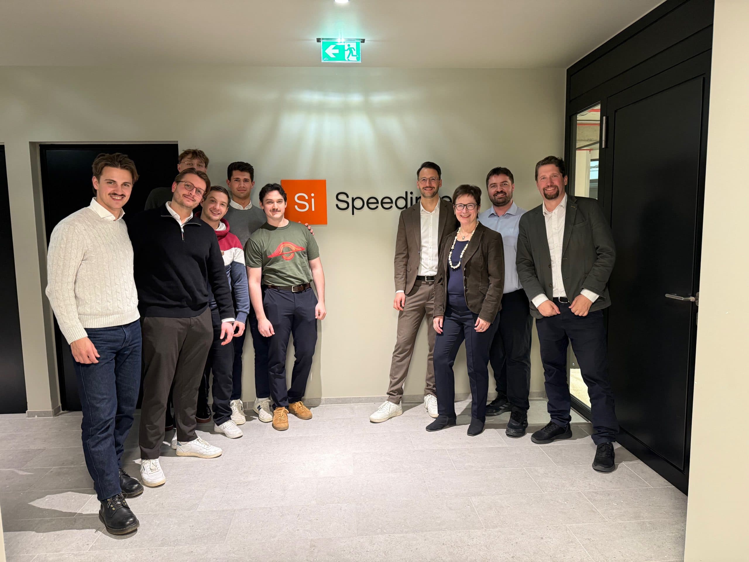 Eine Gruppe von zehn Personen, meist Männer, steht lächelnd in einem Büroflur neben einem Wandschild mit der Aufschrift Si Speed. Sie tragen Business-Casual-Kleidung und stehen unter hellen Deckenleuchten.