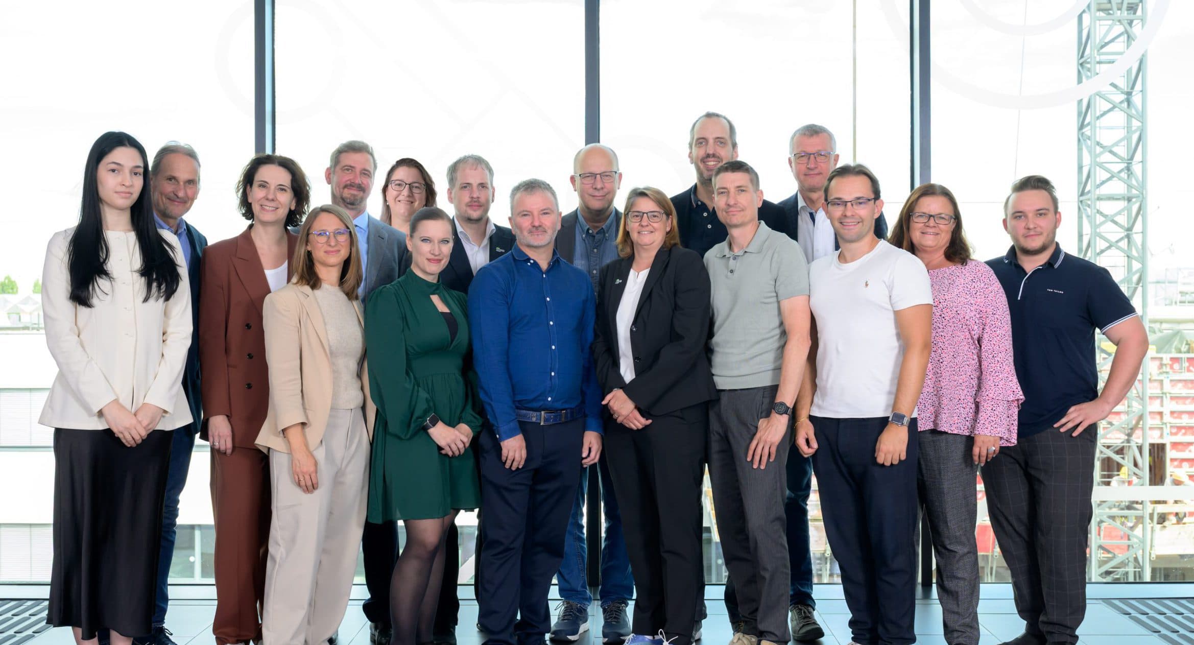 Eine bunt gemischte Gruppe von 19 Personen in legerer Business-Kleidung steht in einem Innenraum vor großen Fenstern mit Blick auf eine Stadtlandschaft und lächelt in die Kamera.