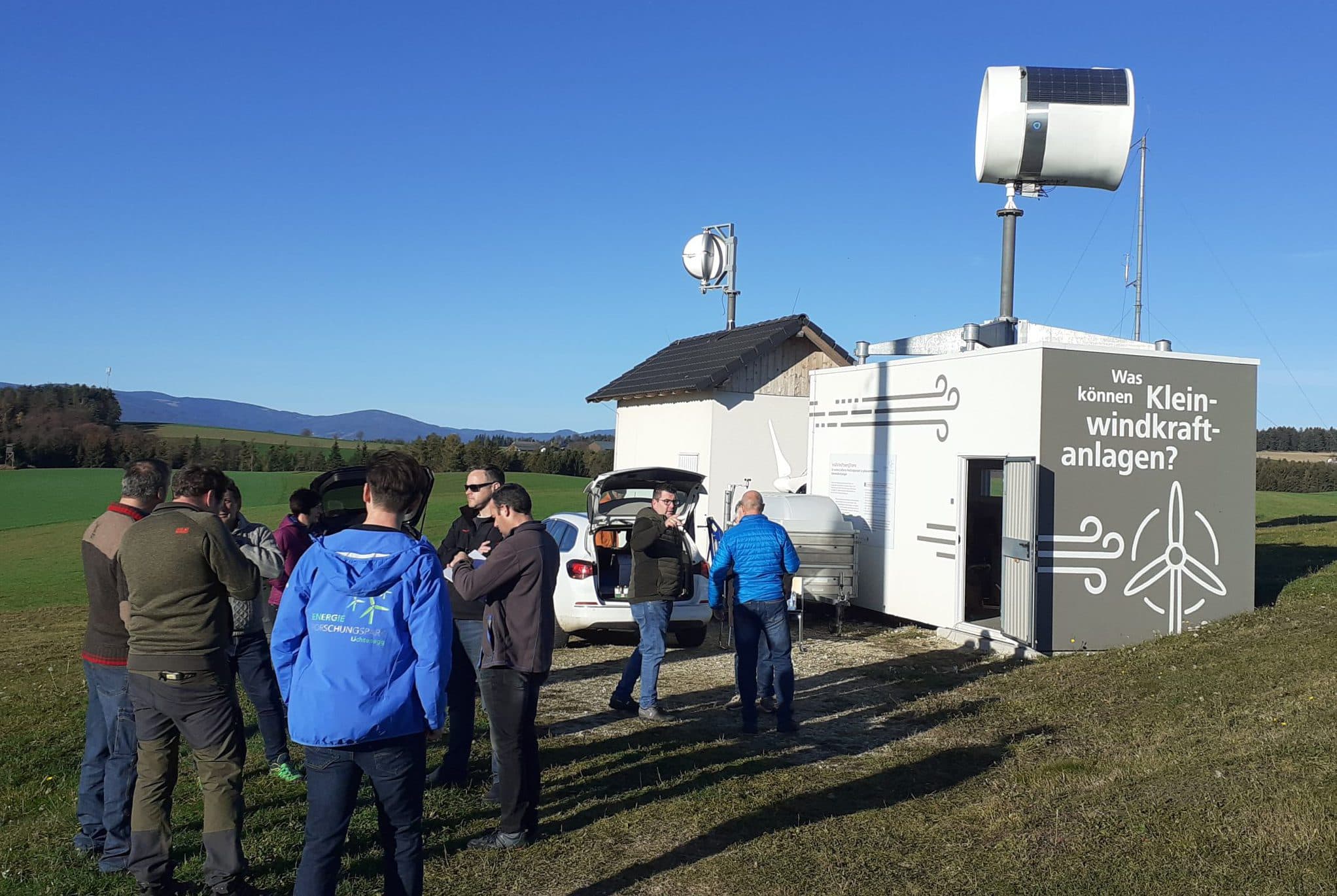Eine Gruppe von Menschen steht in der Nähe einer kleinen Windkraftanlage und eines weißen Gebäudes mit deutscher Aufschrift auf einem grasbewachsenen Feld unter einem klaren blauen Himmel. Im Hintergrund sind sanfte Hügel und Bäume zu sehen.