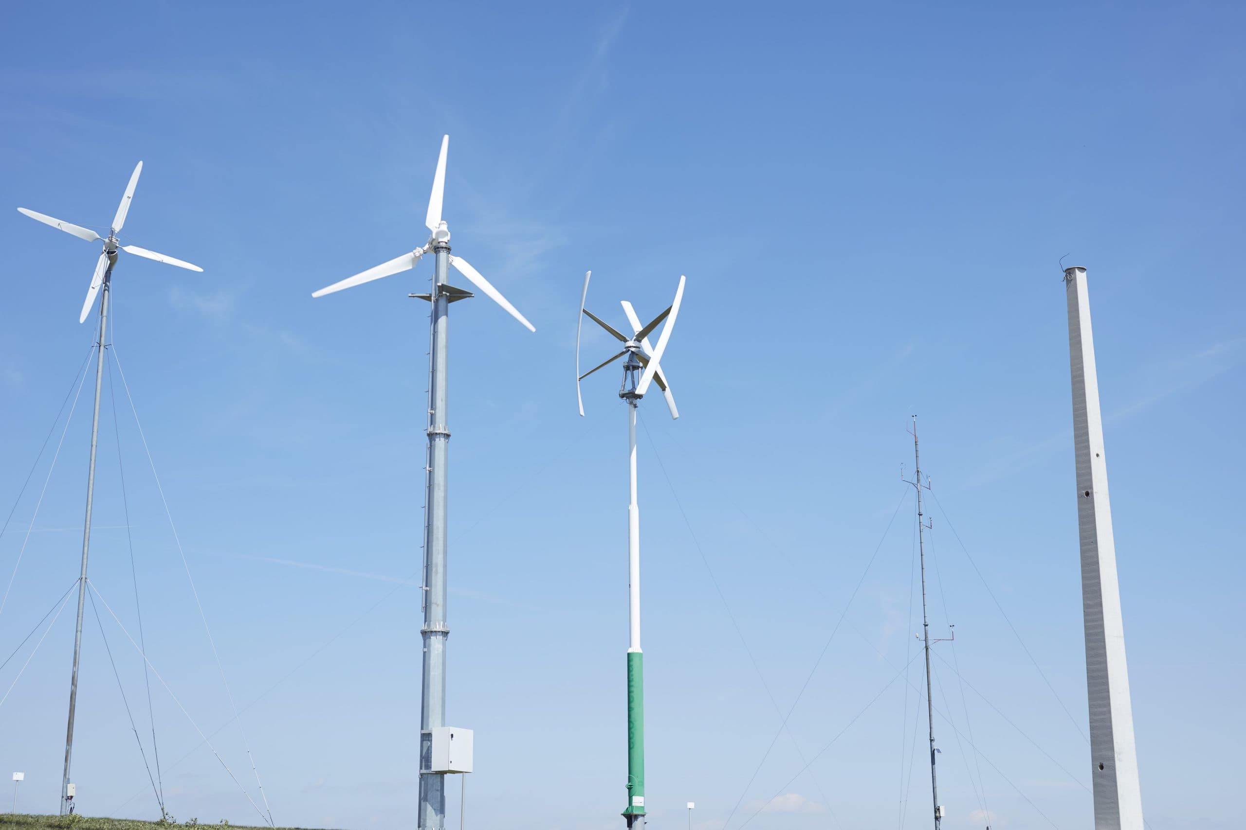 Drei Windturbinen und zwei hohe Masten stehen vor einem strahlend blauen Himmel auf einer Wiese, wobei sich jedes Bauwerk in seiner Form und Höhe unterscheidet.