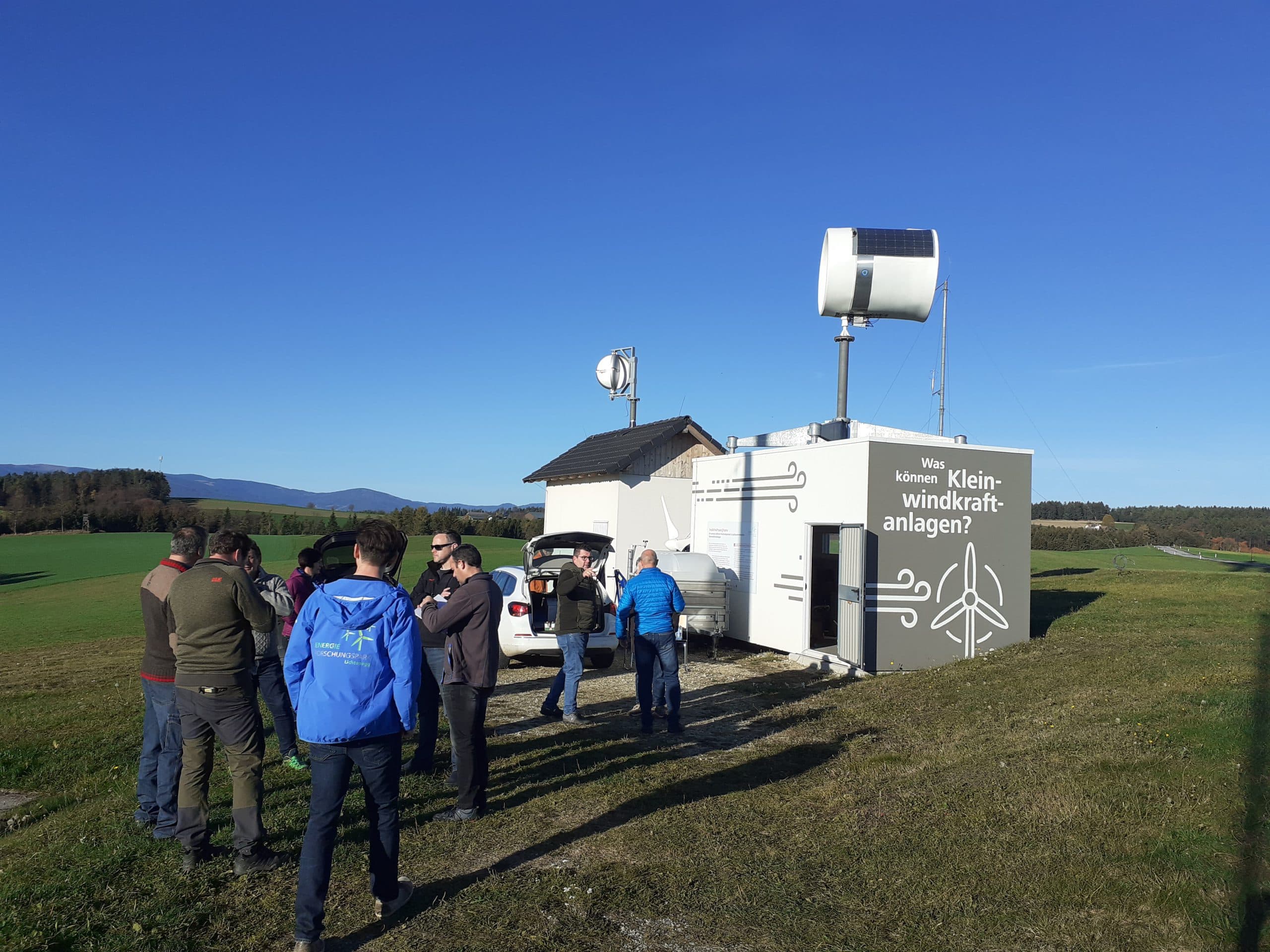 Eine Gruppe von Menschen steht in der Nähe eines kleinen weißen Gebäudes mit einer Windturbine auf dem Dach, das auf einem offenen Feld unter einem klaren blauen Himmel steht. Das Gebäude ist in deutscher Sprache beschriftet und ist von grünem Gras und entfernten Bäumen umgeben.