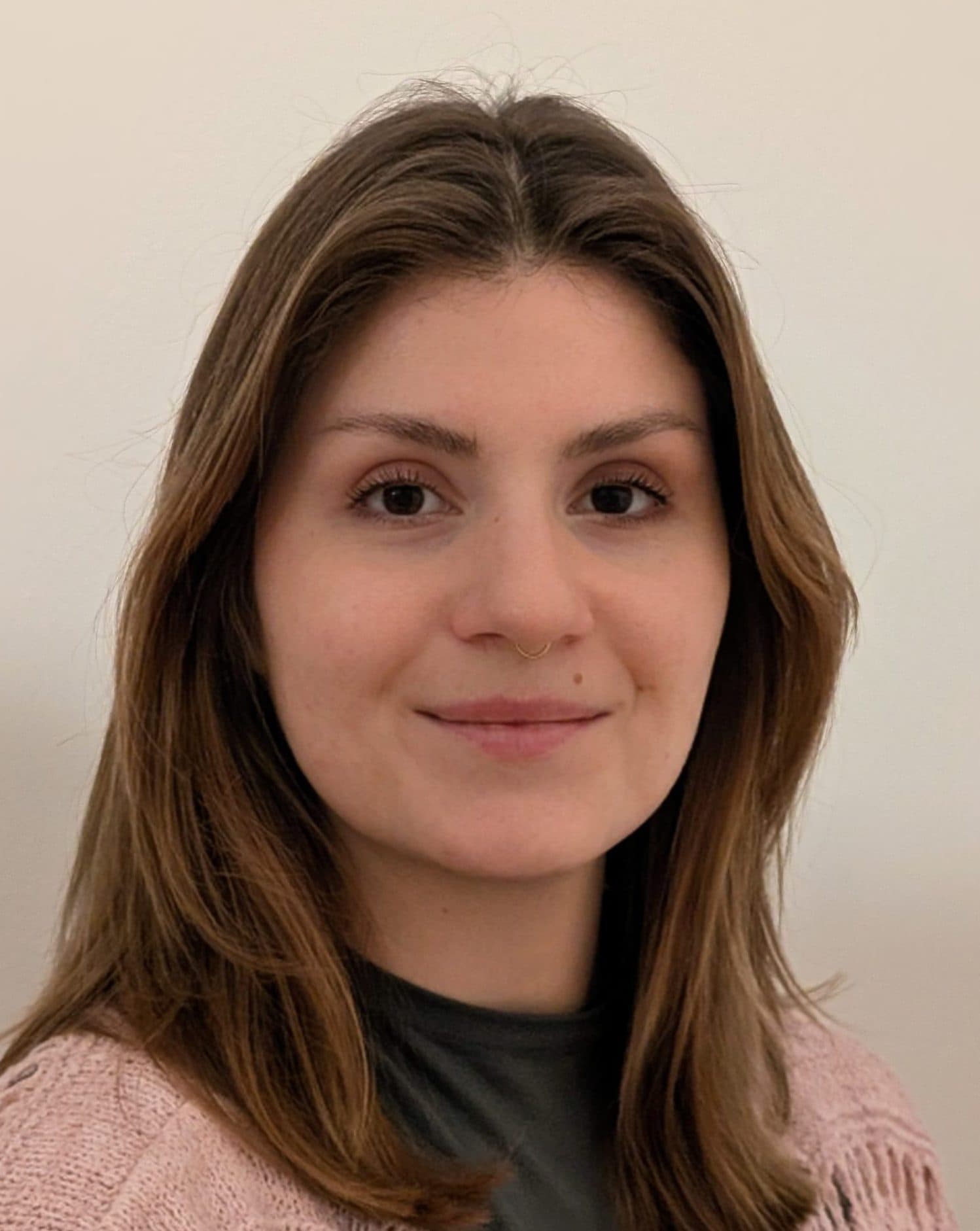 Eine junge Frau mit glattem, schulterlangem braunem Haar lächelt sanft in die Kamera. Sie trägt einen rosa Strickpullover über einem dunklen Oberteil und steht vor einem schlichten, hellen Hintergrund.