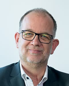 Ein Mann mittleren Alters mit kurzen grauen Haaren, einem Bart und einer Brille trägt eine dunkle Anzugsjacke und ein weißes Hemd. Er lächelt leicht und ist der Kamera zugewandt, mit einem hellen Hintergrund hinter ihm.