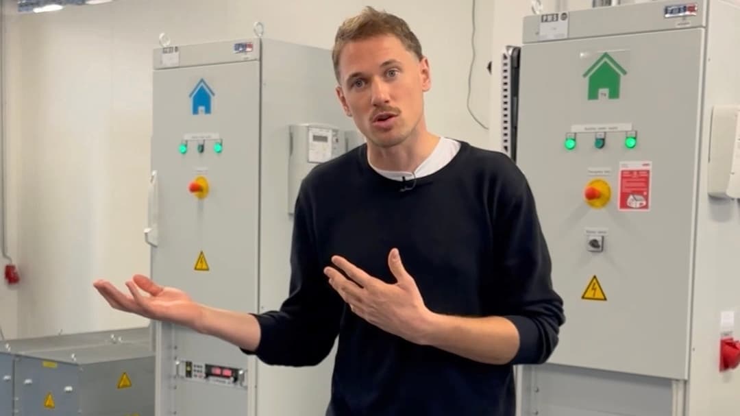 Ein Mann mit kurzen Haaren und schwarzem Pullover gestikuliert mit den Händen, während er vor einer industriellen Schalttafel mit grünen und blauen Haussymbolen und Warnschildern steht.