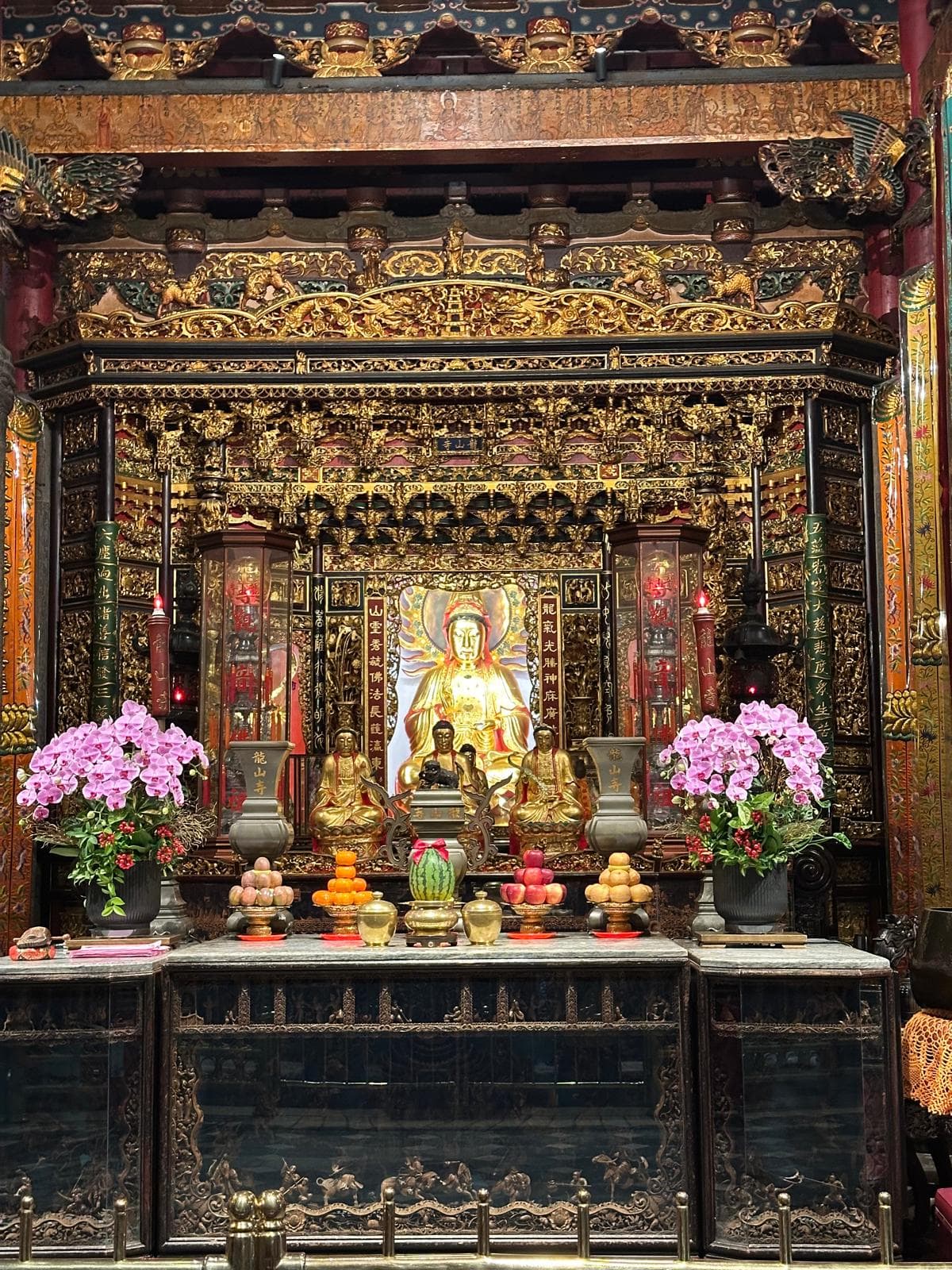 Ein kunstvoller buddhistischer Altar mit goldenen Statuen, leuchtend rosafarbenen Blumenarrangements, Obstopfern und komplizierten dekorativen Schnitzereien in Rot, Gold und Schwarz. Die zentrale Statue ist von kleineren Figuren und Laternen umgeben.