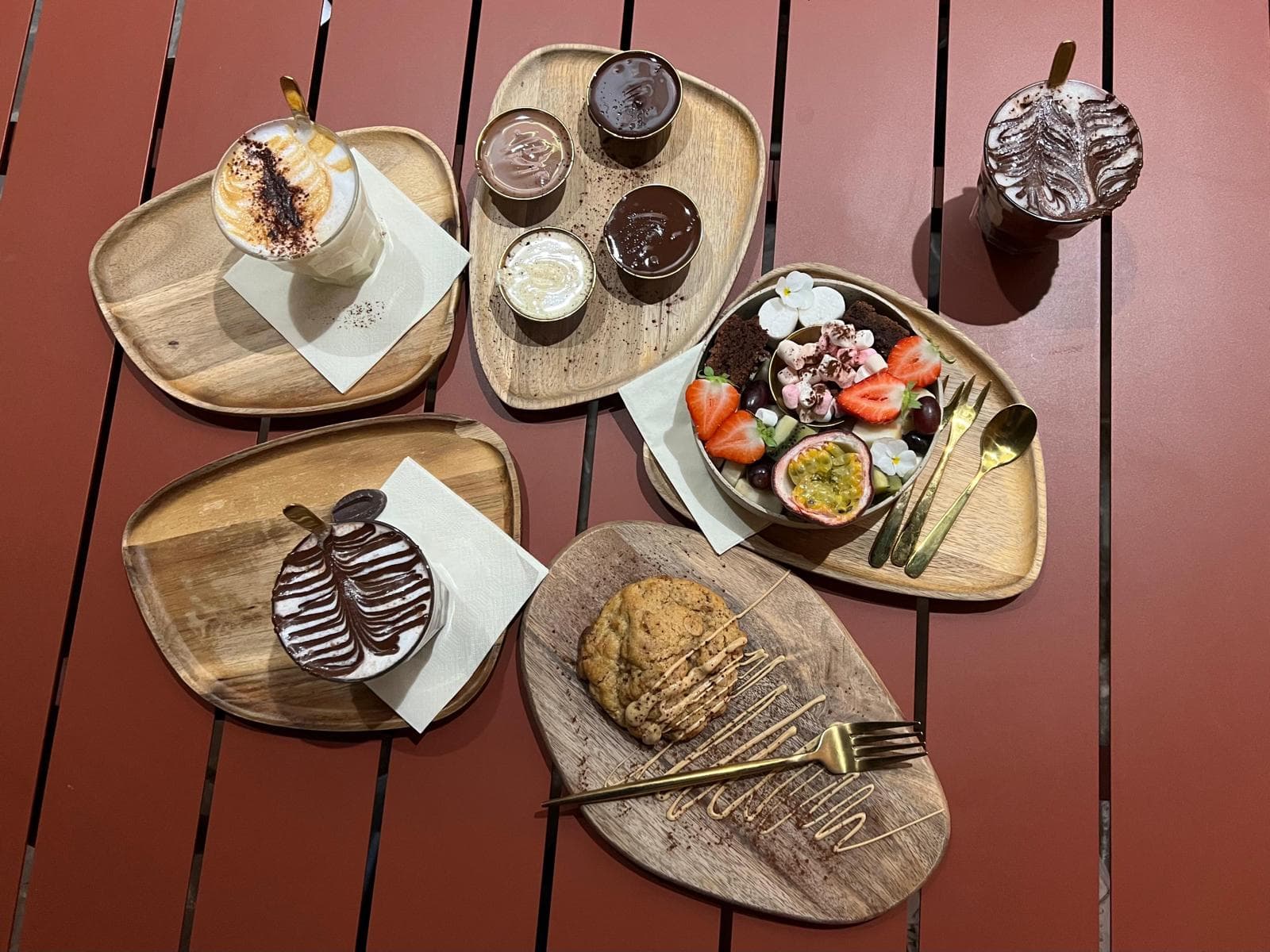 Draufsicht auf einen Tisch mit Holztabletts, auf denen sich verschiedene Desserts befinden, darunter ein Keks, Mousse au Chocolat, ein Getränk mit Schaumkunst, eine Schale mit Obst und Sahne sowie goldfarbene Utensilien, die alle auf einer roten Lattenfläche angeordnet sind.