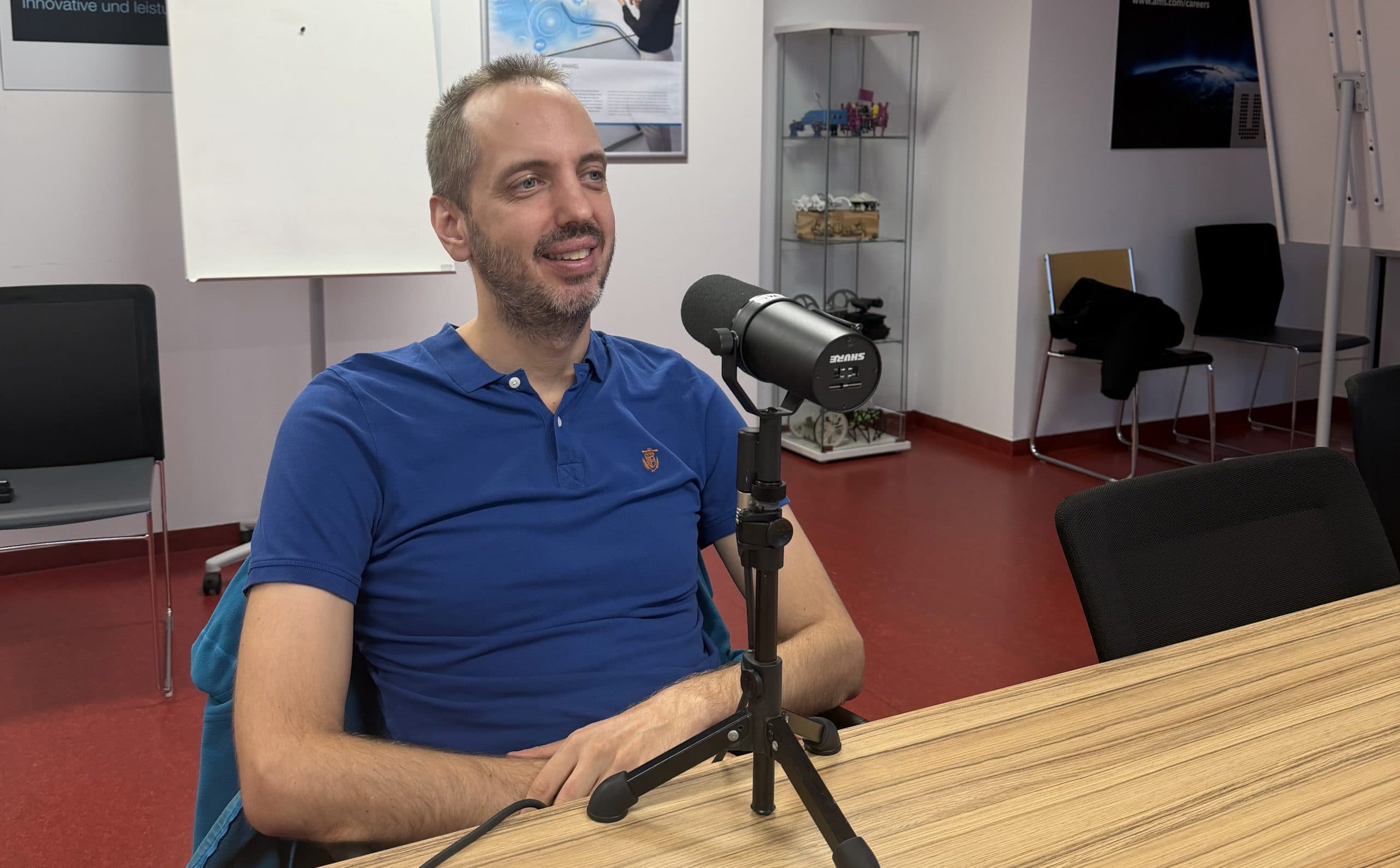 Ein Mann in einem blauen Poloshirt sitzt an einem Tisch in einem modernen Büro, lächelt und spricht in ein Mikrofon auf einem Ständer. Im Hintergrund stehen Stühle, ein Whiteboard und Regale.