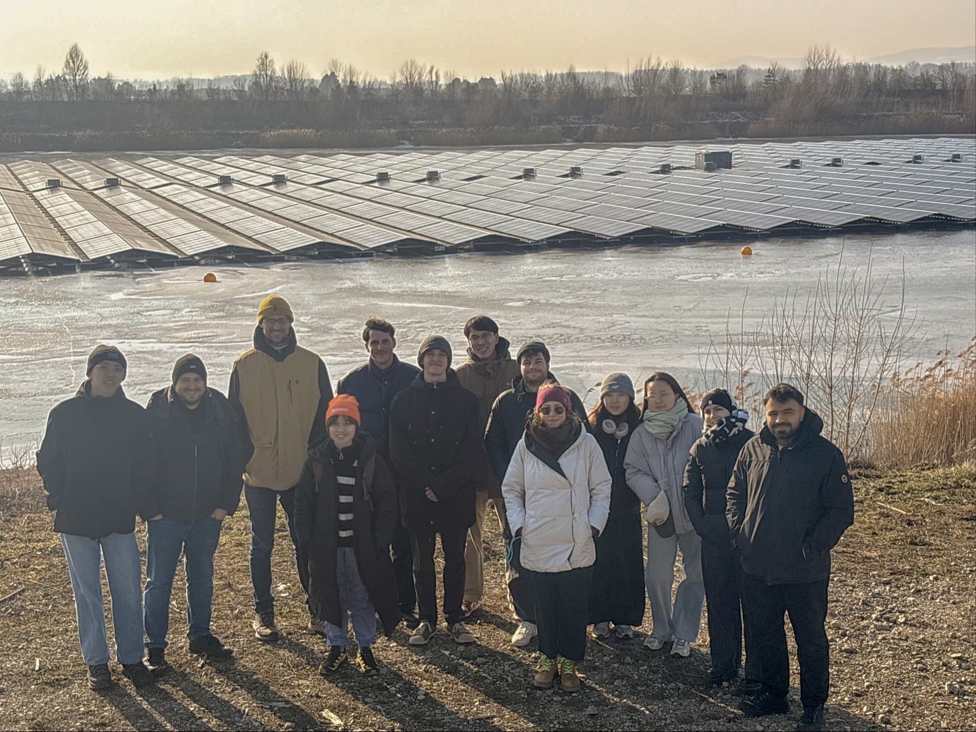 Eine Gruppe von dreizehn Personen steht im Freien, lächelnd in Winterkleidung, vor einer großen Fläche, die an einem sonnigen Tag mit Sonnenkollektoren bedeckt ist. Der Himmel ist dunstig und im Hintergrund sind Bäume zu sehen.