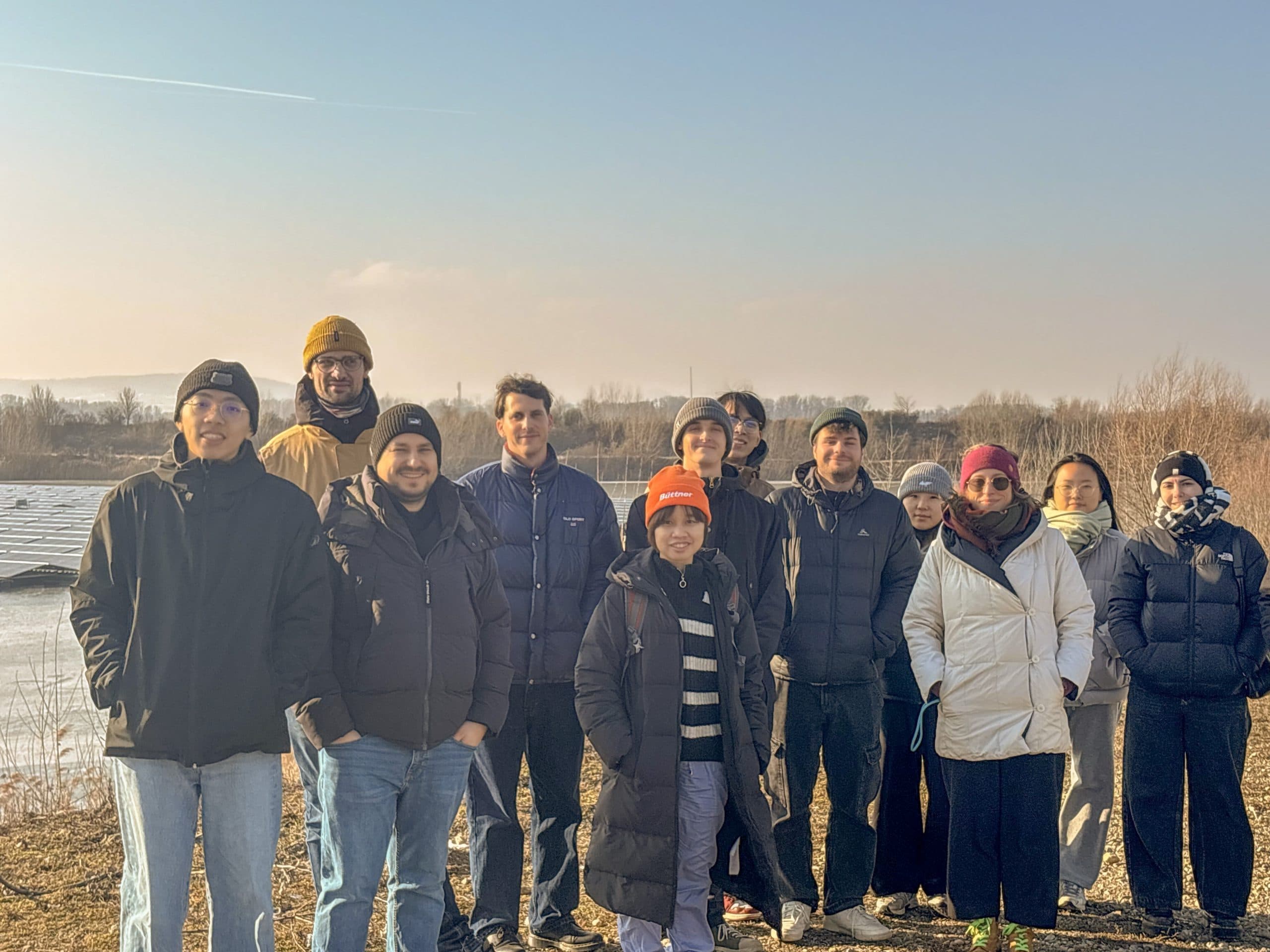 Eine Gruppe von 12 winterlich gekleideten Personen steht im Freien an einem Gewässer mit kahlen Bäumen und klarem Himmel im Hintergrund, lächelt und posiert für ein Foto.