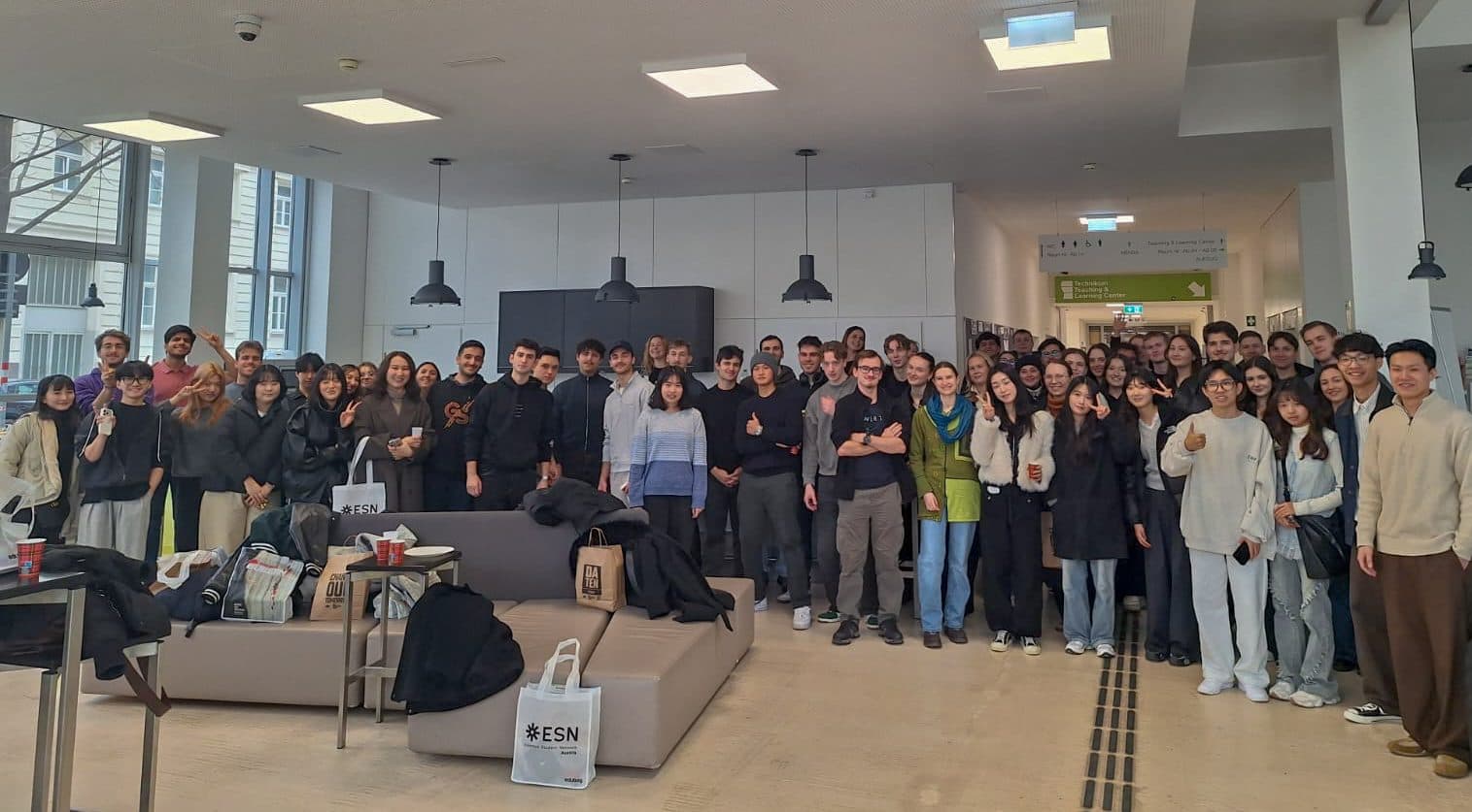 Eine große Gruppe von Menschen, meist junge Erwachsene, steht in einem Innenraum zusammen, lächelt und zeigt die Daumen nach oben. Sie sind leger gekleidet und posieren für ein Gruppenfoto in einem hellen, modernen Raum mit Sofas und Taschen im Vordergrund.