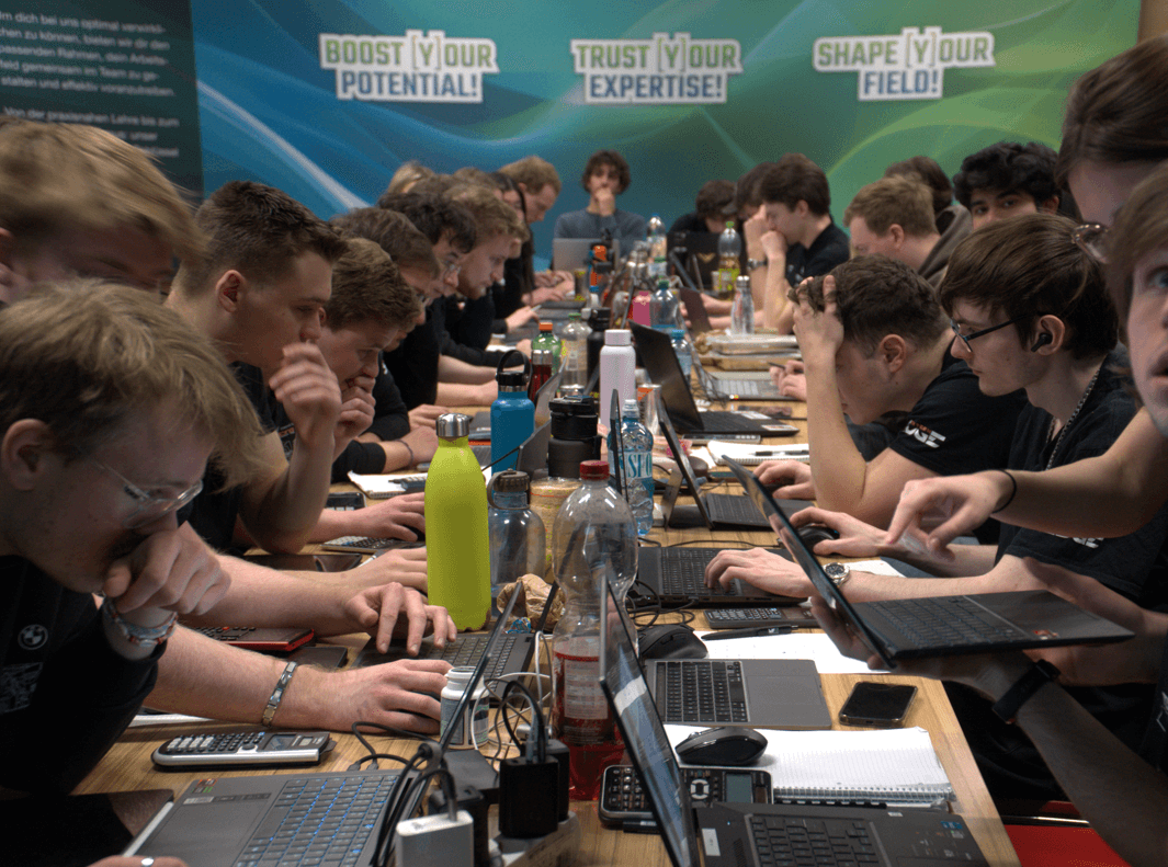 Eine Gruppe junger Leute arbeitet konzentriert an Laptops an einem überfüllten Tisch, mit vielen Wasserflaschen und Schnüren, unter motivierenden Schildern, auf denen "Boost Your Potential!", "Trust Your Expertise!" und "Shape Your Field!" steht.