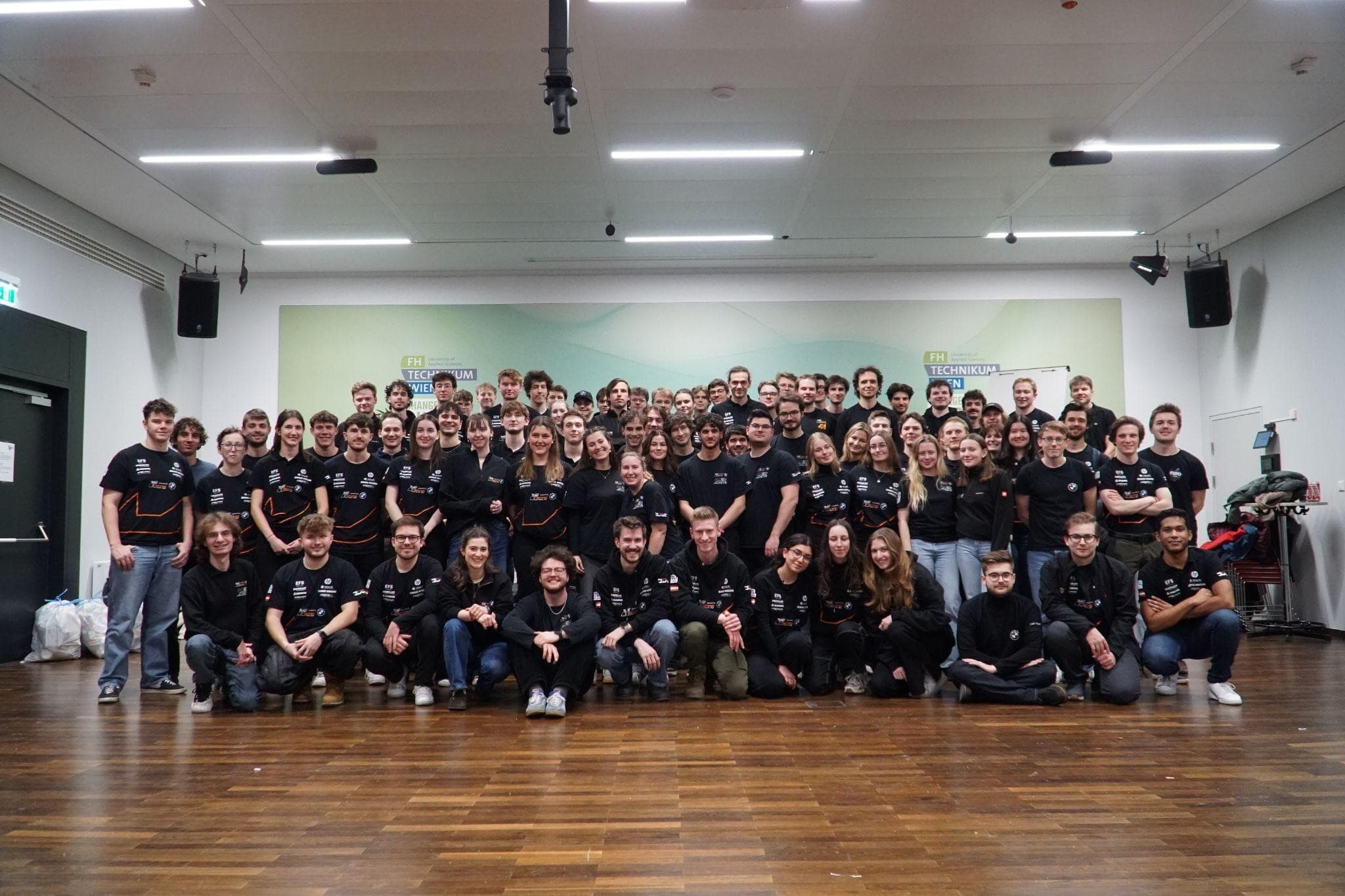 Eine große Gruppe von Menschen in passenden schwarzen Hemden posiert in einem Innenraum und lächelt in die Kamera. Sie stehen und knien auf einem Holzboden in einem hellen Raum mit einer grün-weißen Fahne im Hintergrund.