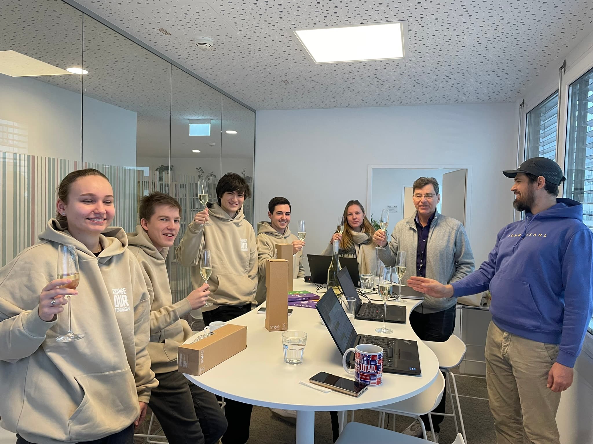 Sechs Personen stehen um einen Tisch in einem Büro, lächeln und halten Gläser mit Champagner in der Hand. Laptops und Kisten stehen auf dem Tisch, und die Gruppe scheint gemeinsam einen Anlass zu feiern.