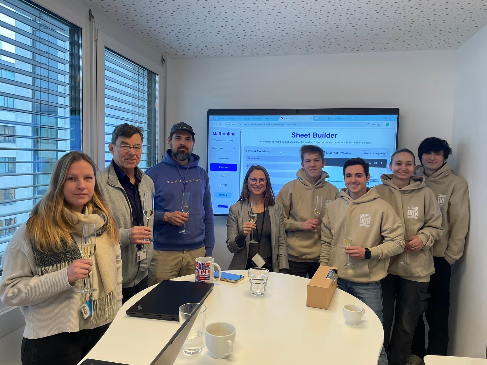 Sieben Personen stehen in einem Besprechungsraum, halten Gläser in der Hand und lächeln, während hinter ihnen ein großer Bildschirm mit einer Sheet Builder-App angezeigt wird. Auf dem Tisch vor ihnen stehen Tassen, Becher und ein Laptop.