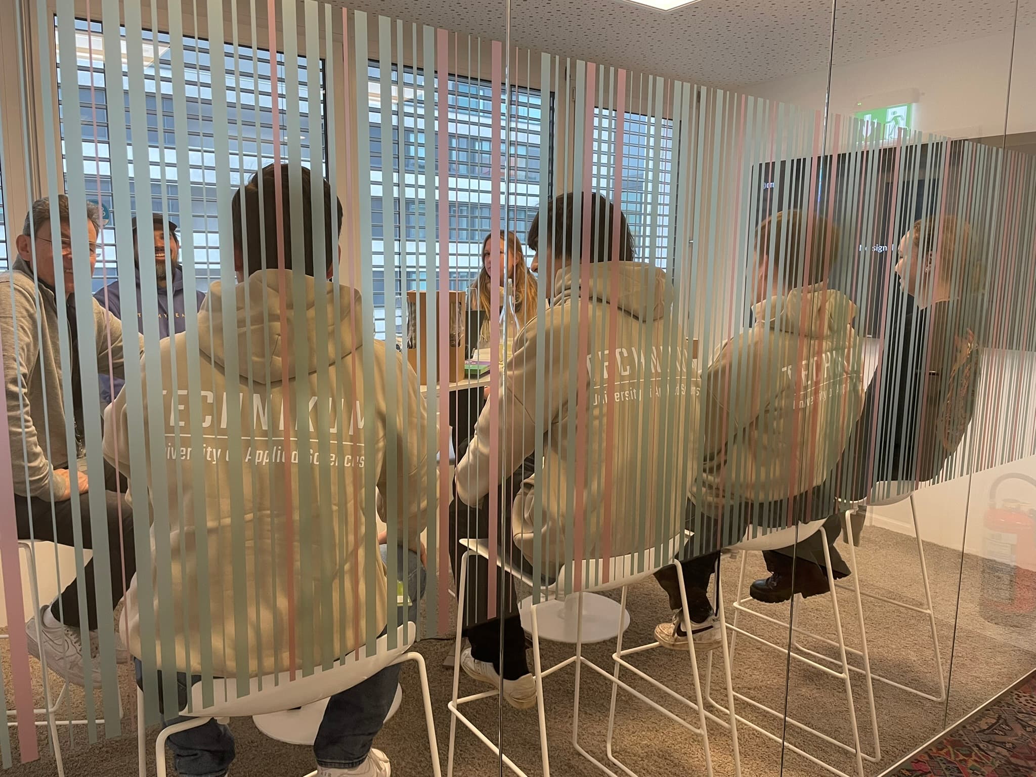Eine Gruppe von Menschen sitzt auf Hockern in einem gläsernen Besprechungsraum, der durch vertikale Streifen auf dem Glas zu sehen ist. Einige tragen passende Kapuzenpullis und ein paar Leute sind in ein Gespräch vertieft.