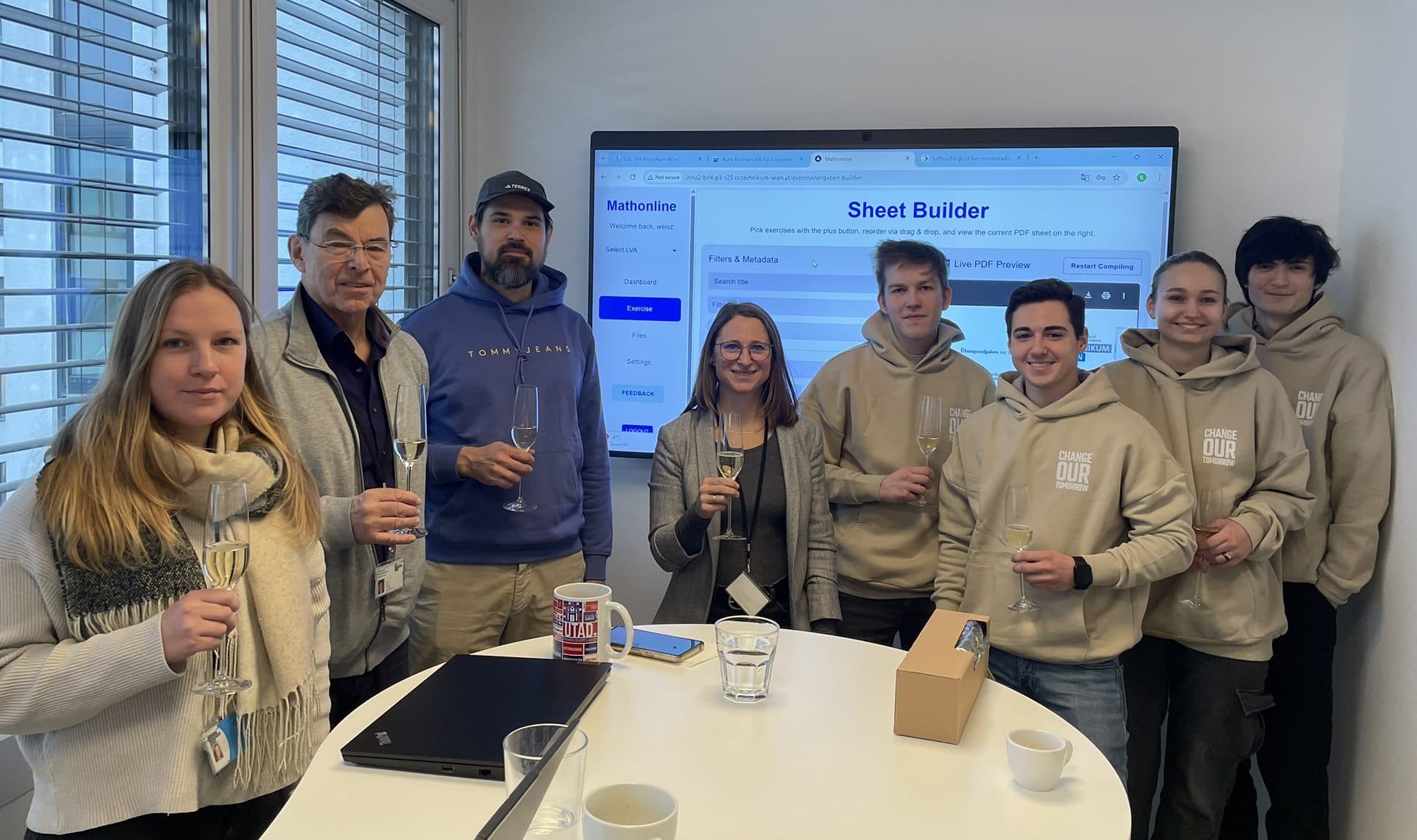 Sieben Personen, einige in passenden Kapuzenpullovern, stehen in einem modernen Büro um einen Tisch mit Getränken, Tassen und Laptops. Hinter ihnen befindet sich ein großer Bildschirm mit einer Sheet Builder-Webseite. Sie posieren und lächeln in die Kamera.