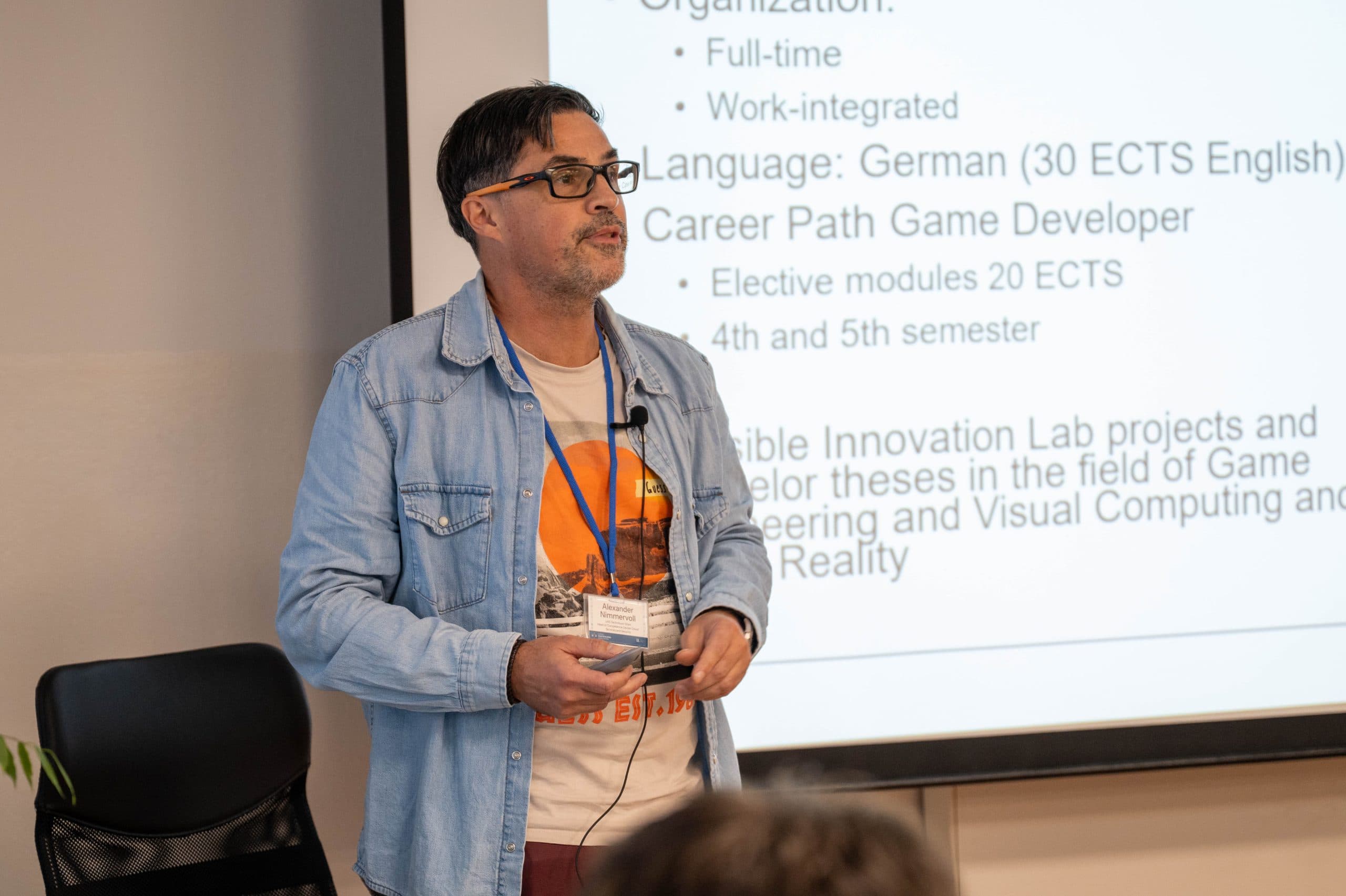 Ein Mann, der eine Brille, ein Jeanshemd und ein orangefarbenes Schlüsselband trägt, steht vor einer projizierten Präsentationsfolie und spricht zu einem Publikum in einem Klassenzimmer oder Seminar.
