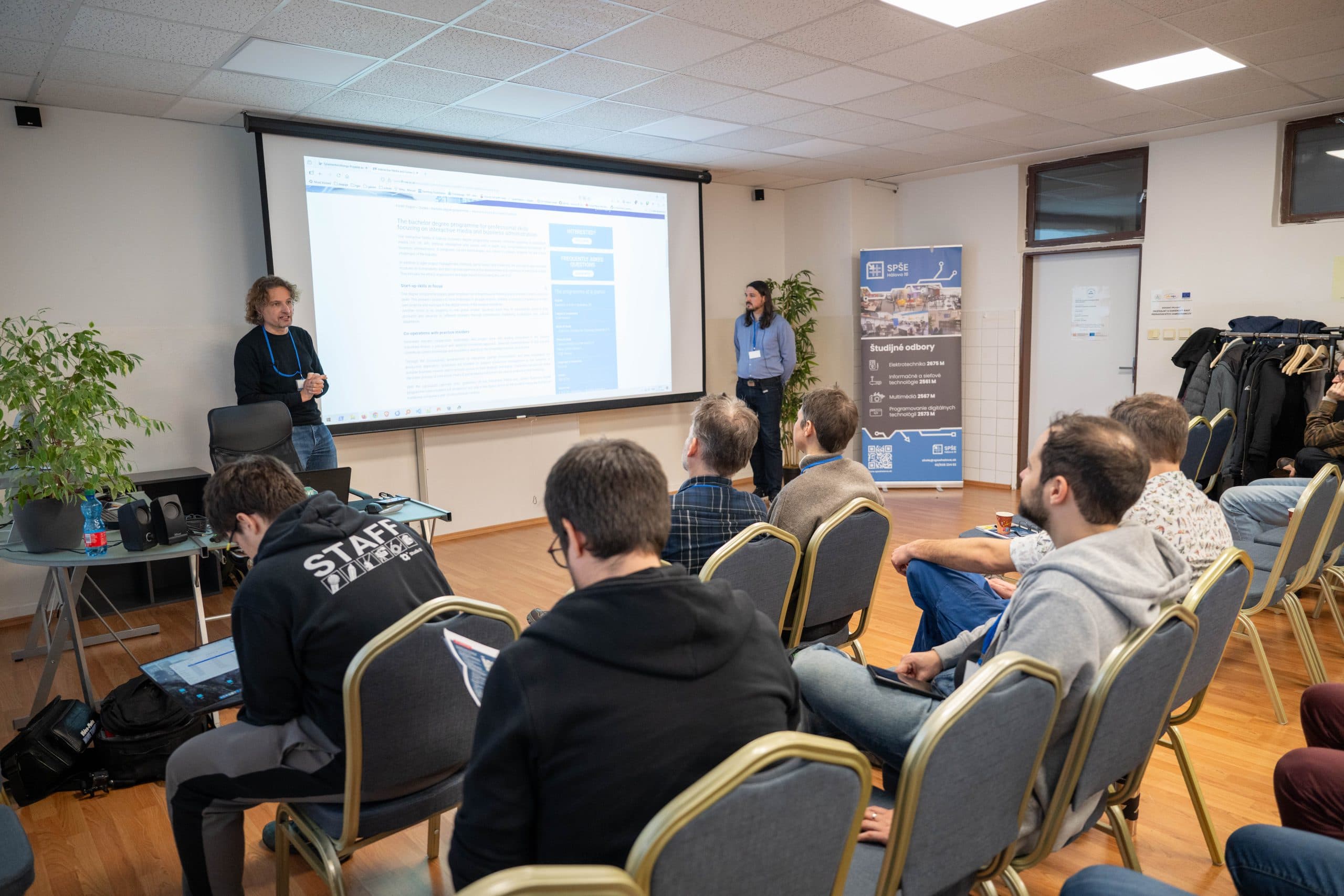 Eine Gruppe von Personen sitzt auf Stühlen vor einem Projektor in einem Konferenzraum. Zwei Vortragende stehen vorne, einer spricht, während die Präsentationsfolien auf dem Bildschirm angezeigt werden. Einige Teilnehmer verwenden Laptops.