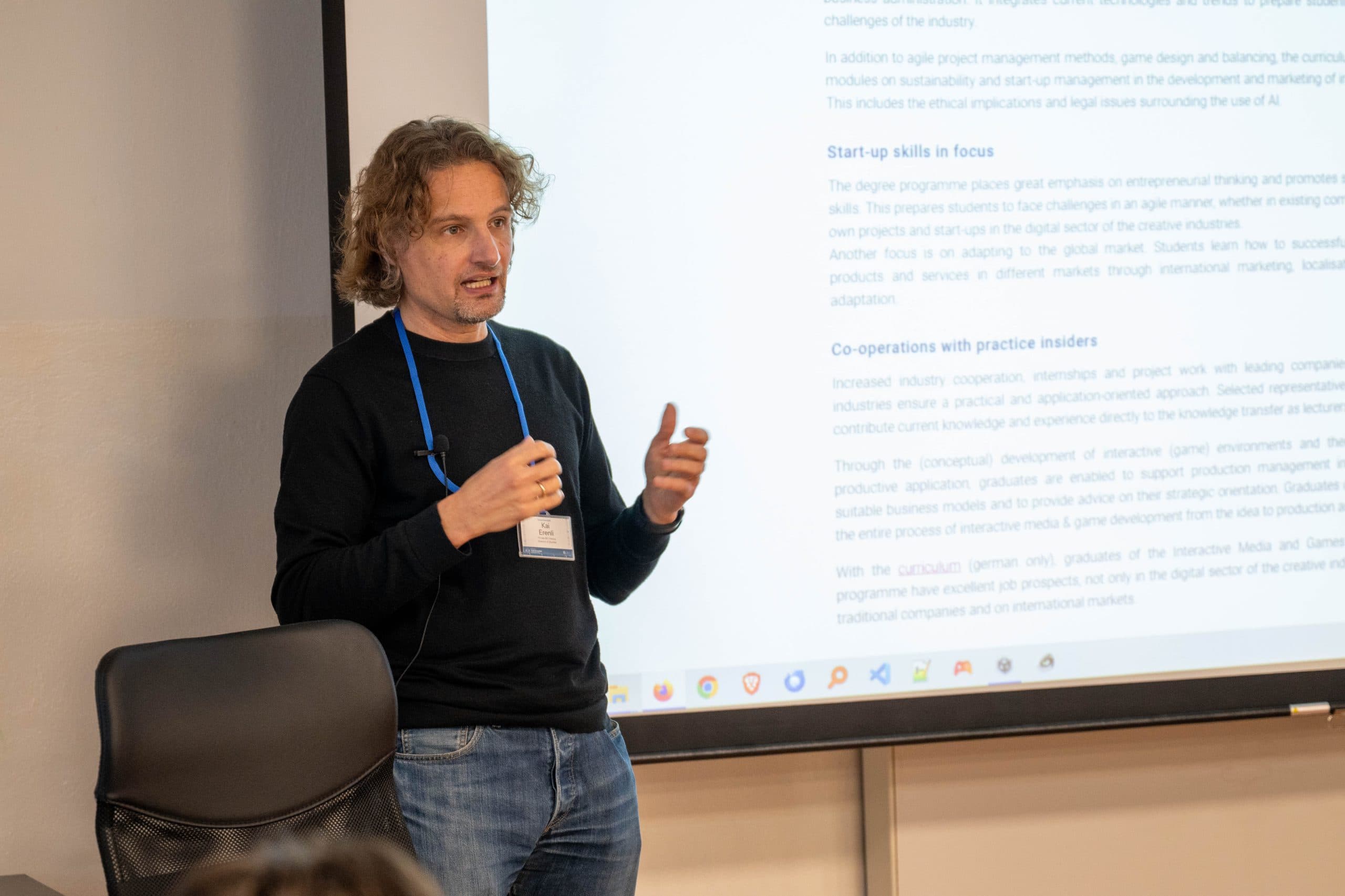 Ein Mann mit lockigem Haar, schwarzem Pullover und blauem Schlüsselband steht vor einer Projektionsfläche, auf der Text während einer Präsentation angezeigt wird, und spricht.