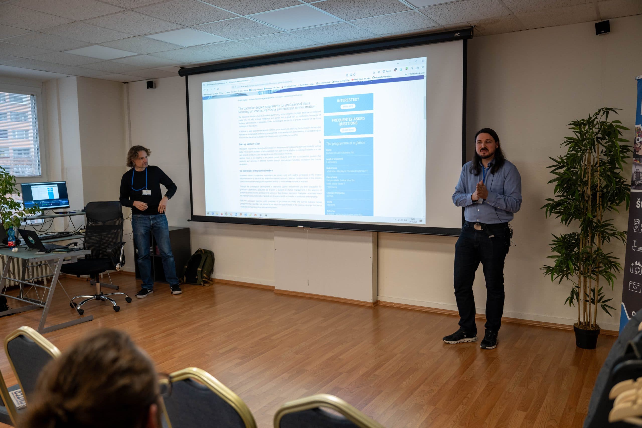 Zwei Personen stehen vor einem Projektor und halten eine Präsentation in einem Konferenzraum. Einer steht an einem Schreibtisch mit Monitoren, während der andere zu einem Publikum spricht, das in Stuhlreihen sitzt.