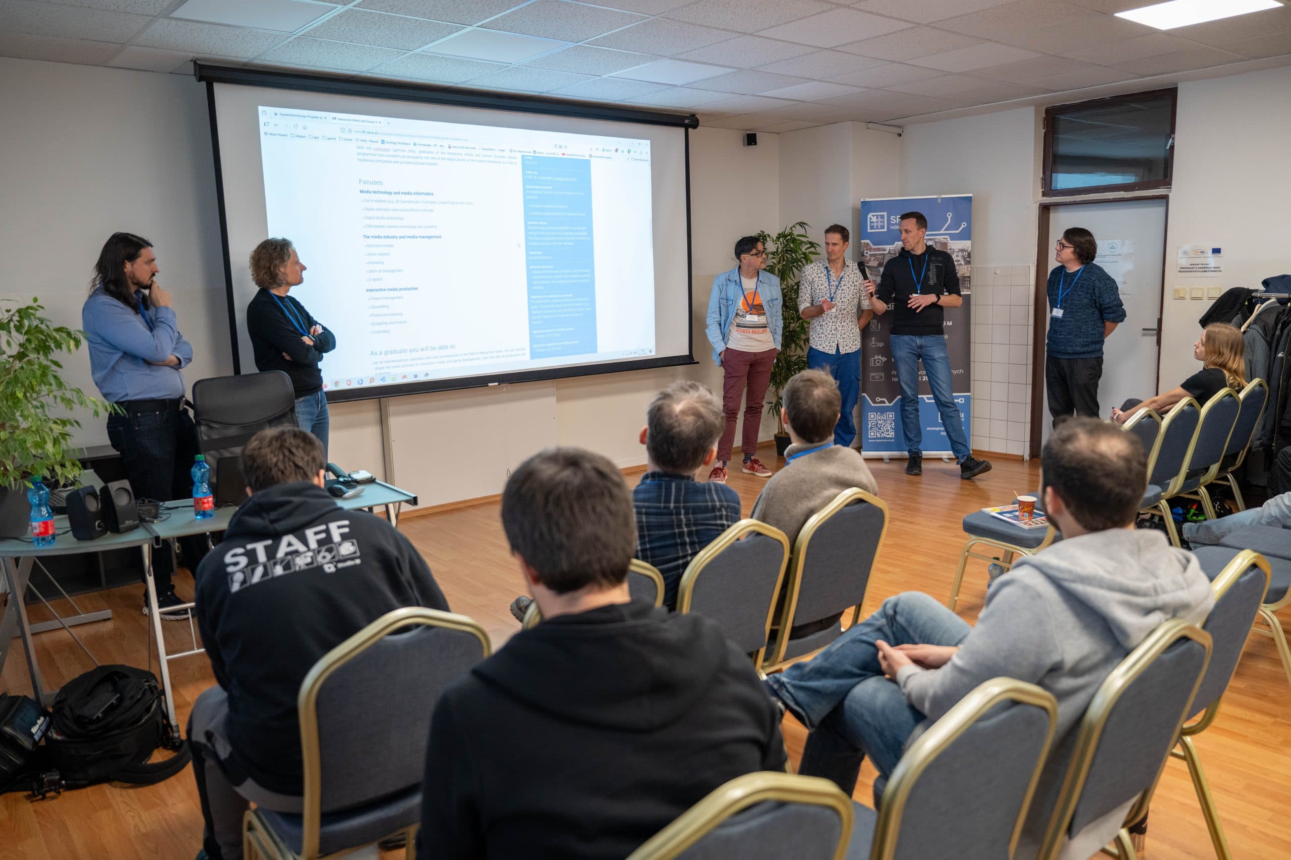 Eine Gruppe von Personen steht vorne in einem Konferenzraum und hält eine Präsentation vor einem Publikum, das auf Stühlen sitzt. Auf einem großen Bildschirm wird ein Text an der Wand hinter ihnen angezeigt. Einige Teilnehmer tragen Namensschilder und Mitarbeiterhemden.