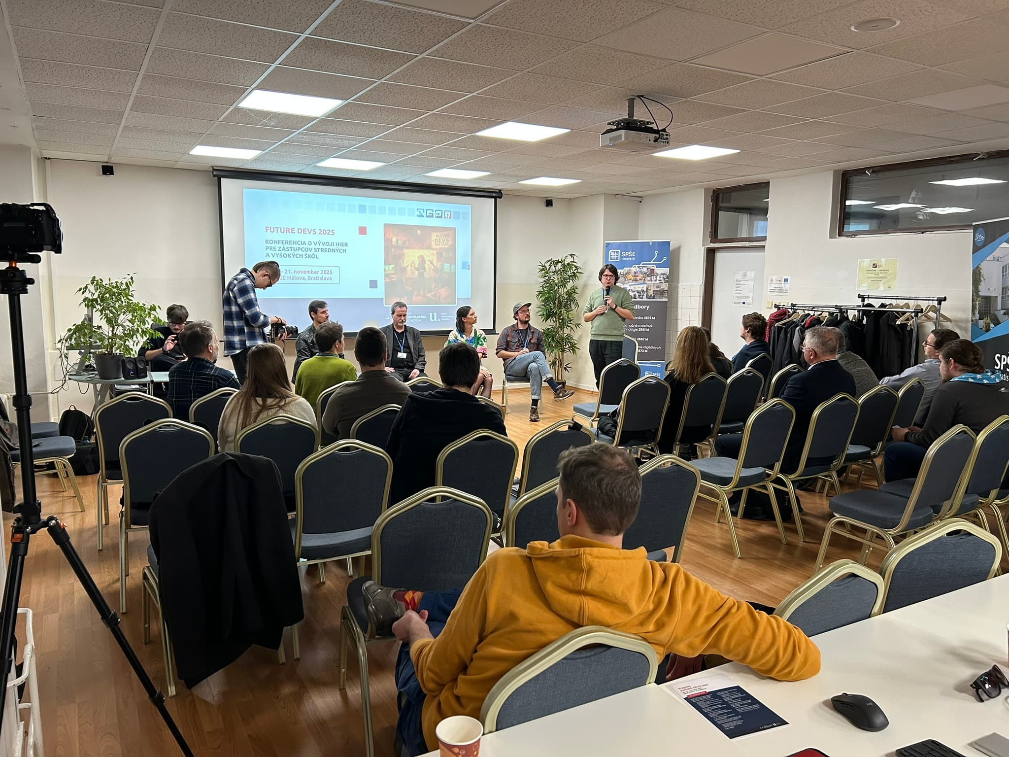 Eine Gruppe von Personen sitzt in Reihen vor einer Gruppe von Rednern im vorderen Teil eines Konferenzraums. Eine Präsentation wird auf eine Leinwand projiziert, und ein Redner steht, während die anderen sitzen. Einige Teilnehmer machen sich Notizen.