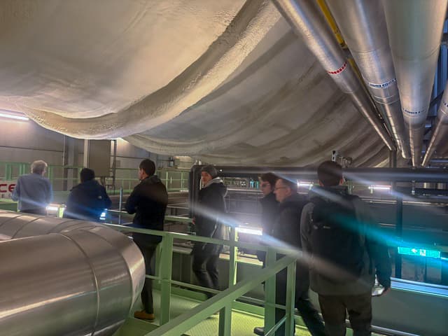Eine Gruppe von dunkel gekleideten Personen geht über einen Metallweg in einer Industrieanlage mit großen Rohren und einer gewölbten, isolierten Decke über dem Kopf. Helle Lichter erhellen den Bereich.
