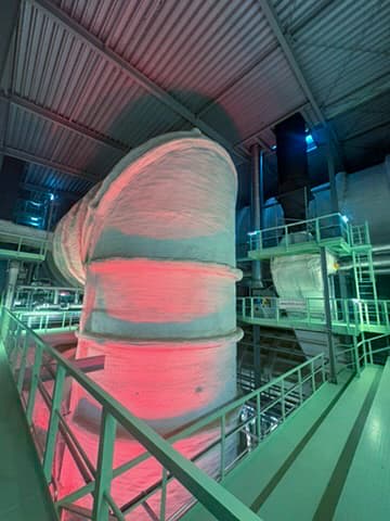 Ein großes, weiß isoliertes Industrierohr steht in einer Fabrik mit grünen Geländern und Plattformen, die von roten und blauen Lichtern beleuchtet werden, unter einer hohen Metalldecke.