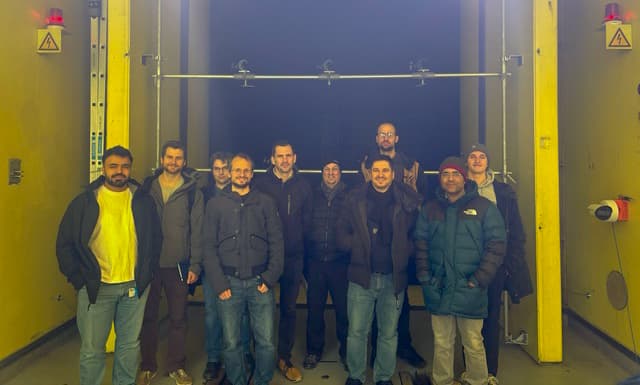 Eine Gruppe von elf Personen in Winterjacken steht zusammen und lächelt vor einer großen gelben Industrieanlage mit schwarzen und gelben Wänden.