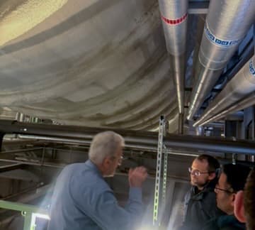Drei Personen stehen in einer industriellen Umgebung unter großen isolierten Rohren und Tanks und besprechen oder überprüfen die Geräte über ihnen. Die Beleuchtung ist gedämpft und die Umgebung wirkt technisch.