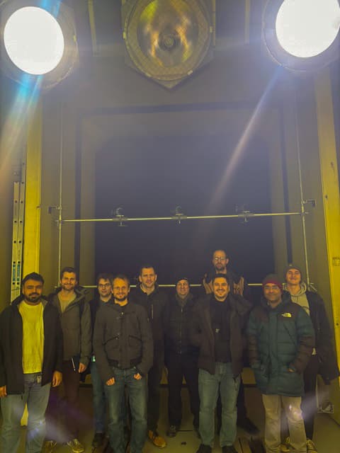 Eine Gruppe von zehn Personen steht lächelnd in zwei Reihen in einer Industriehalle, die von zwei großen Oberlichtern beleuchtet wird. Alle tragen warme Jacken und Freizeitkleidung.