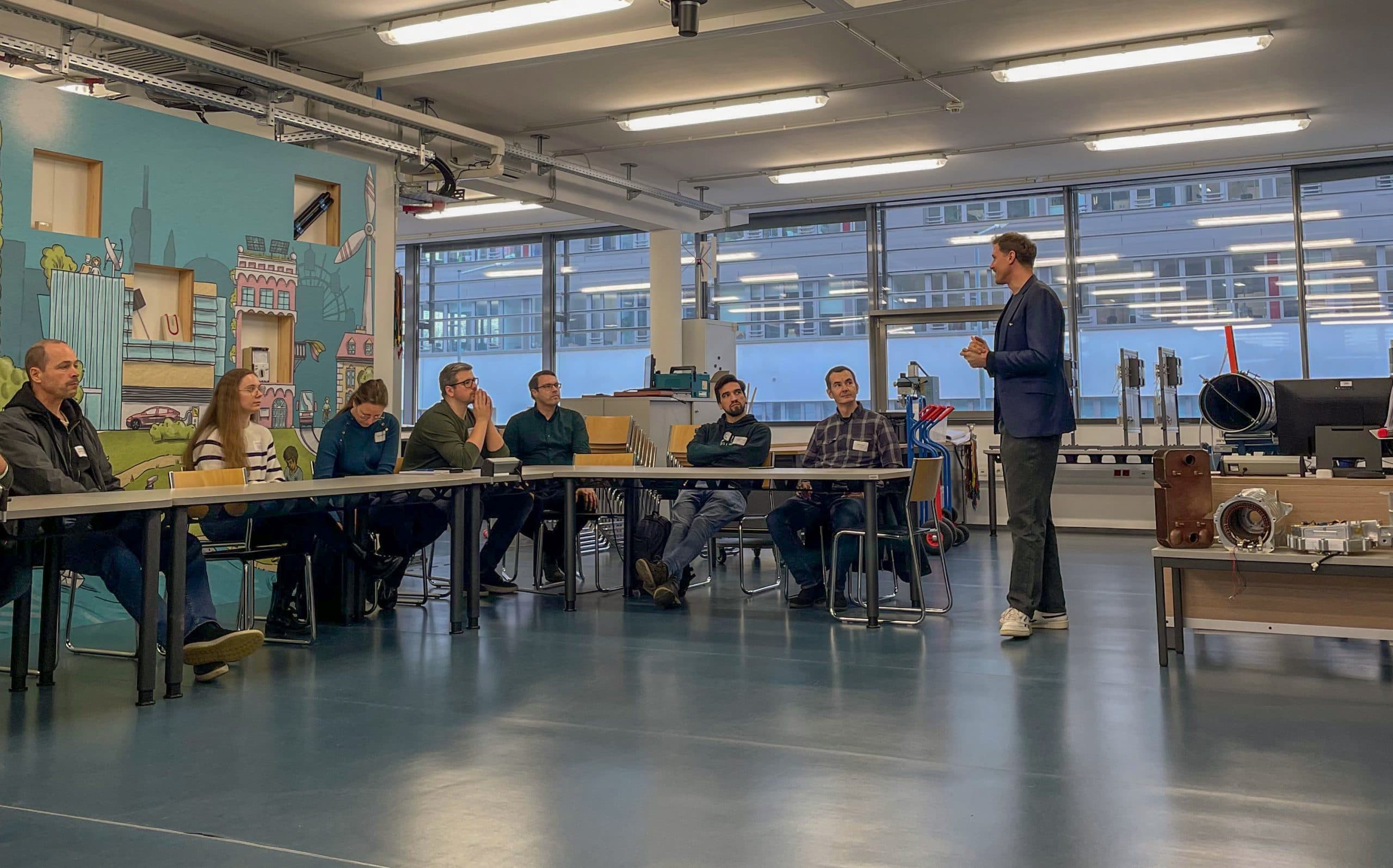 Ein Mann in einem blauen Blazer steht auf und spricht zu einer Gruppe von Menschen, die an Tischen in einem hellen, modernen Klassenzimmer mit großen Fenstern und farbenfroher Wandkunst sitzen.