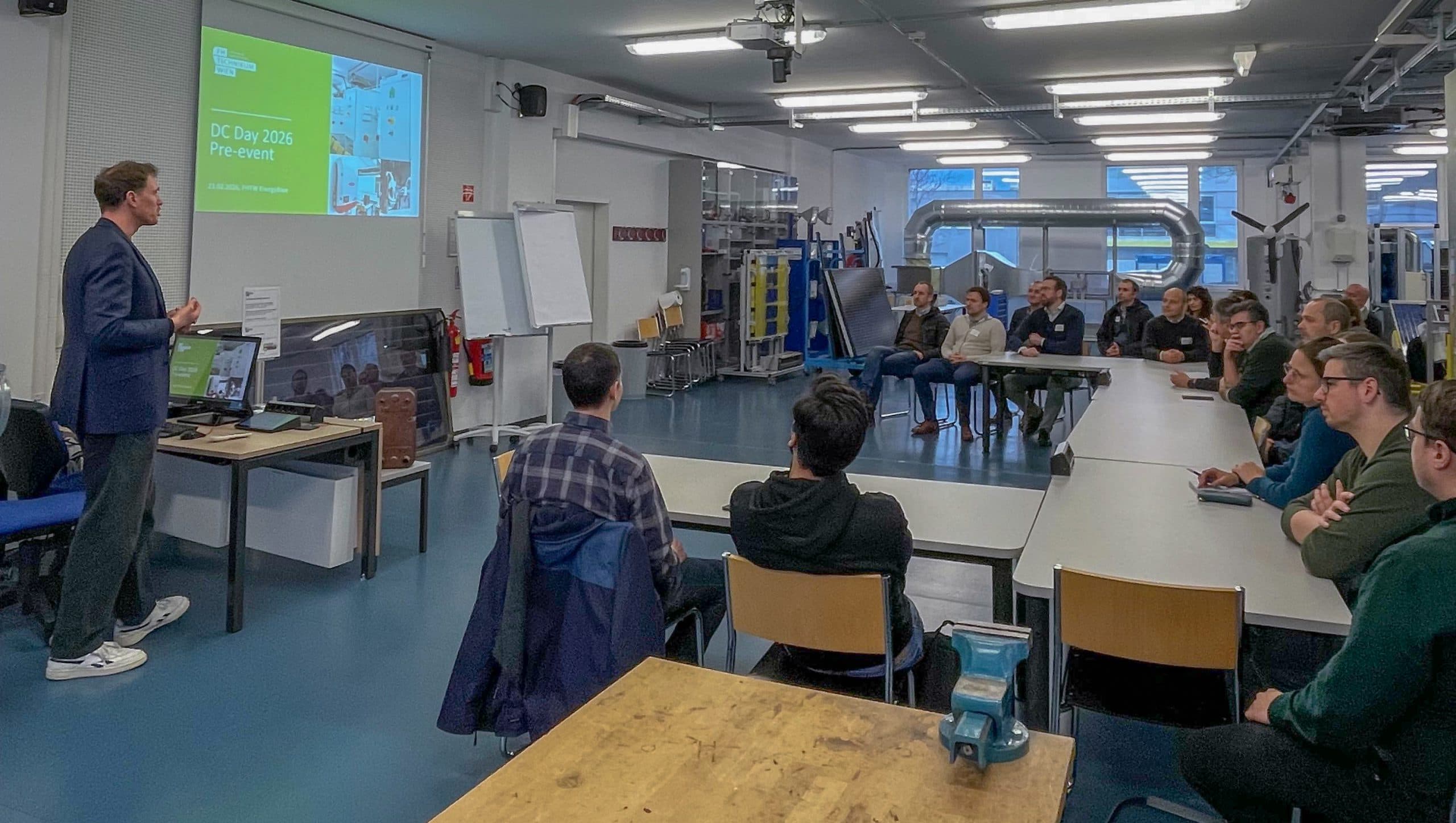 Ein Mann steht vor einer Gruppe von Menschen, die in einem modernen Klassenzimmer an Tischen sitzen. Auf einem großen Bildschirm werden der DC Day 2016 Pre-Event und ein Diagramm angezeigt. Alle scheinen engagiert und aufmerksam zu sein.