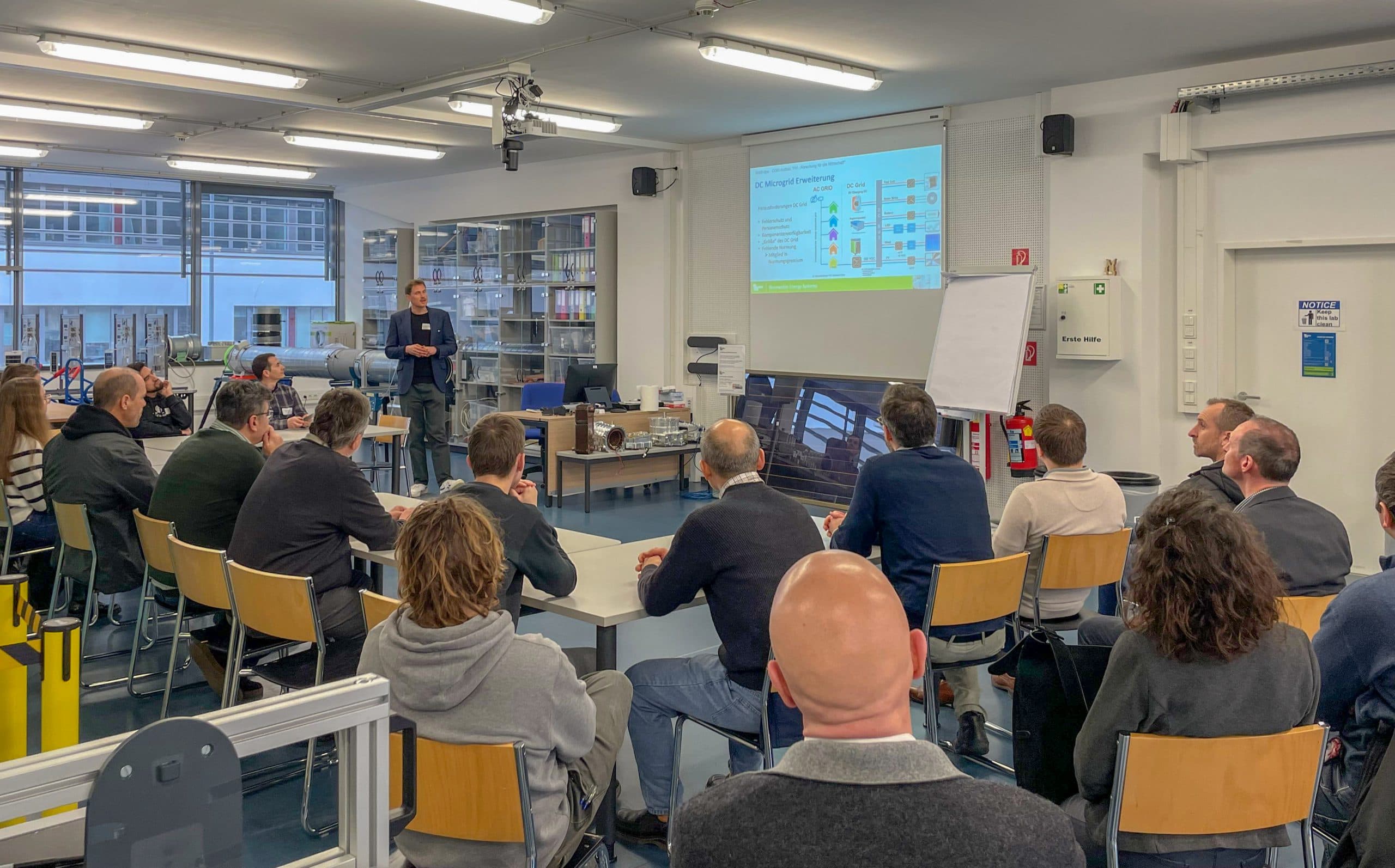 Eine Gruppe von Personen sitzt in einem Klassenzimmer und blickt auf einen Vortragenden, der vor einem Bildschirm spricht, auf dem eine bunte Präsentation gezeigt wird. Der Raum ist hell, mit großen Fenstern und Regalen im Hintergrund.