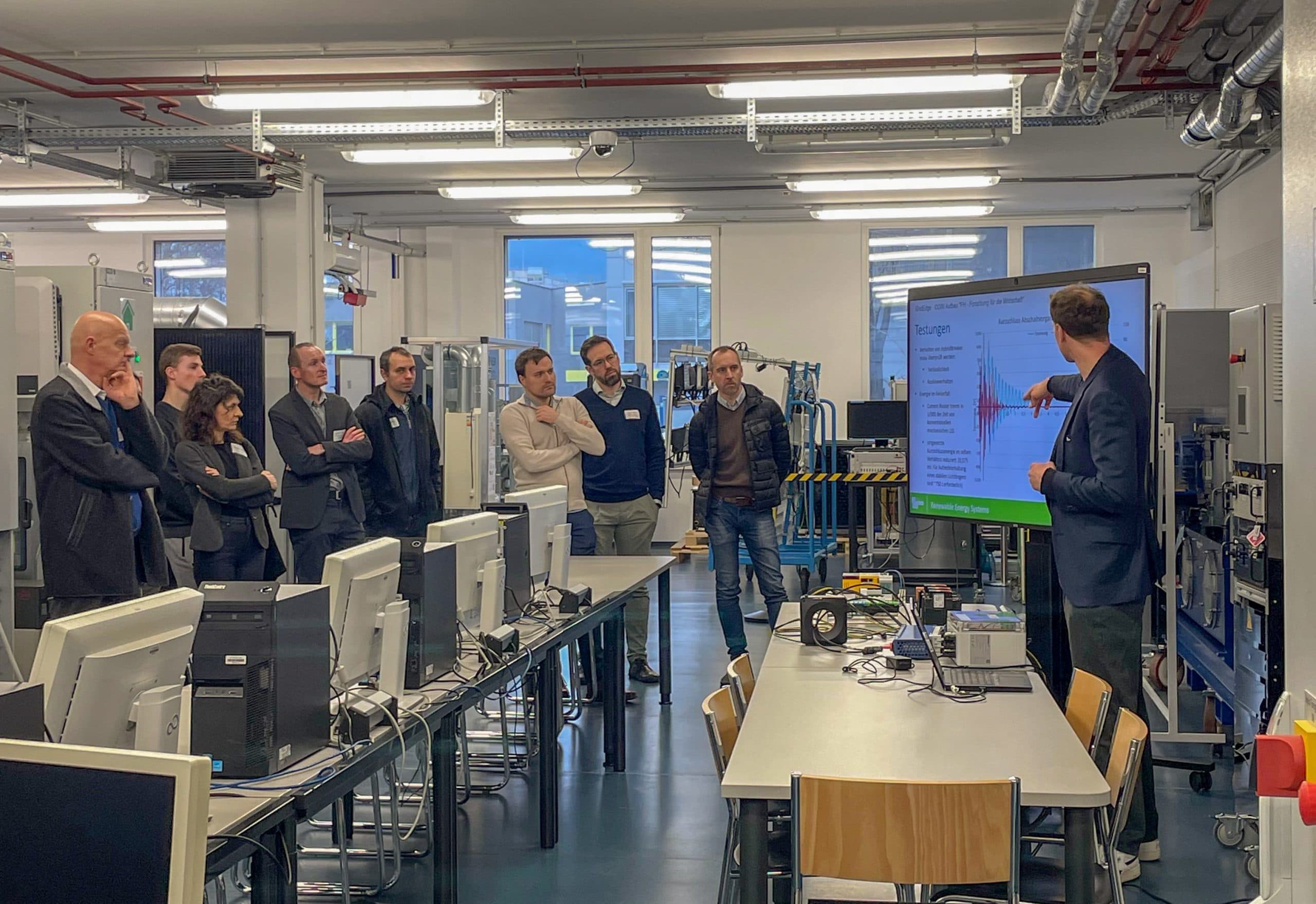 Eine Gruppe von Personen in Geschäftskleidung steht in einem modernen Labor und hört aufmerksam einem Mann zu, der Informationen auf einem großen Bildschirm präsentiert. Der Raum ist mit Computern, Schreibtischen und Laborgeräten ausgestattet.