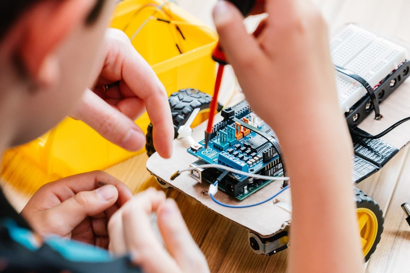 Nahaufnahme von Händen, die eine elektronische Schaltung an einem kleinen Roboterauto mit gelben Rädern montieren, indem sie einen Schraubenzieher benutzen und Drähte an eine Leiterplatte auf einem Holztisch anschließen.