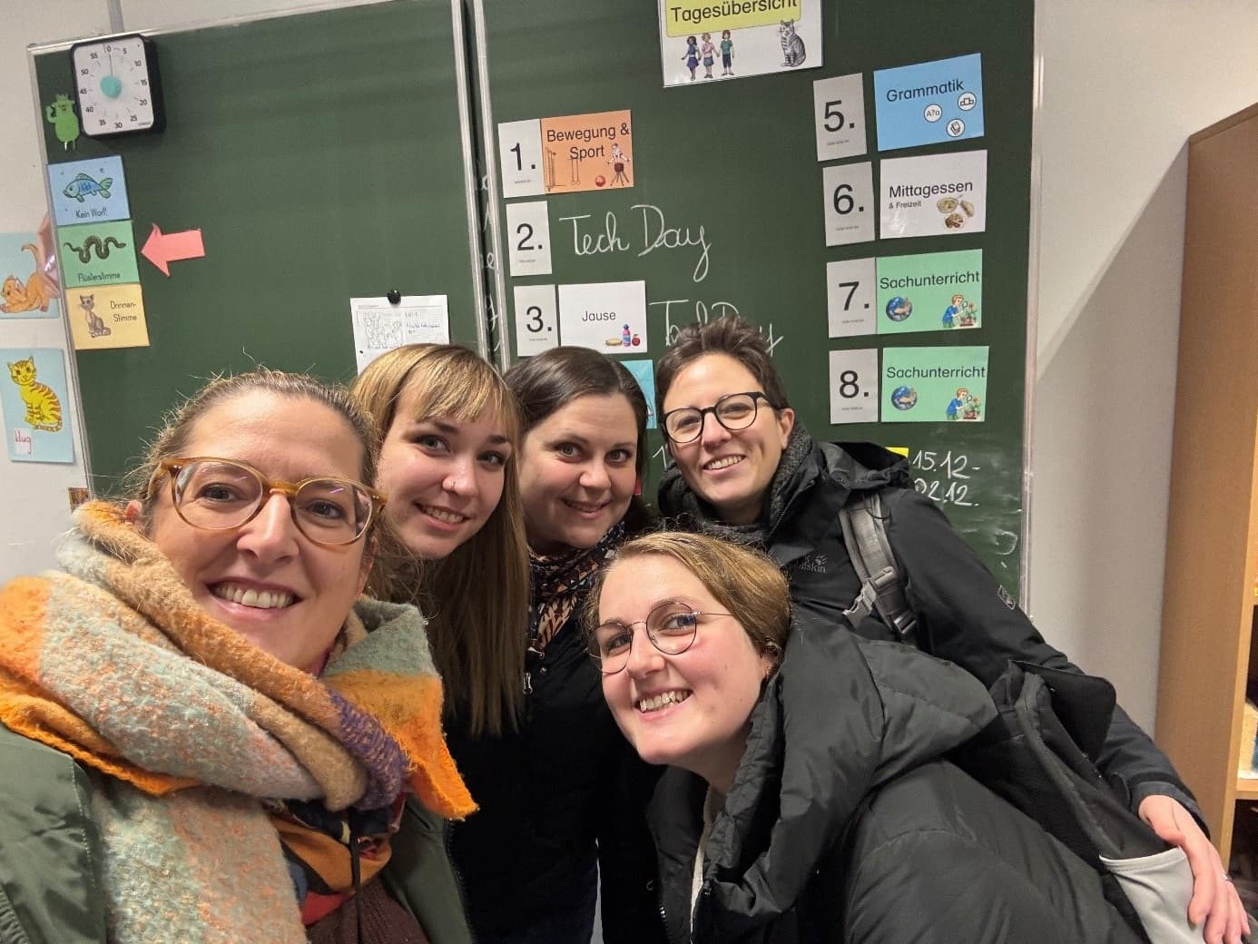 Fünf lächelnde Frauen posieren in einem Klassenzimmer vor einer Kreidetafel, auf der ein Stundenplan und der Tag der Technik in englischer und deutscher Sprache stehen. An der Wand sind bunte Bildungsposter zu sehen.