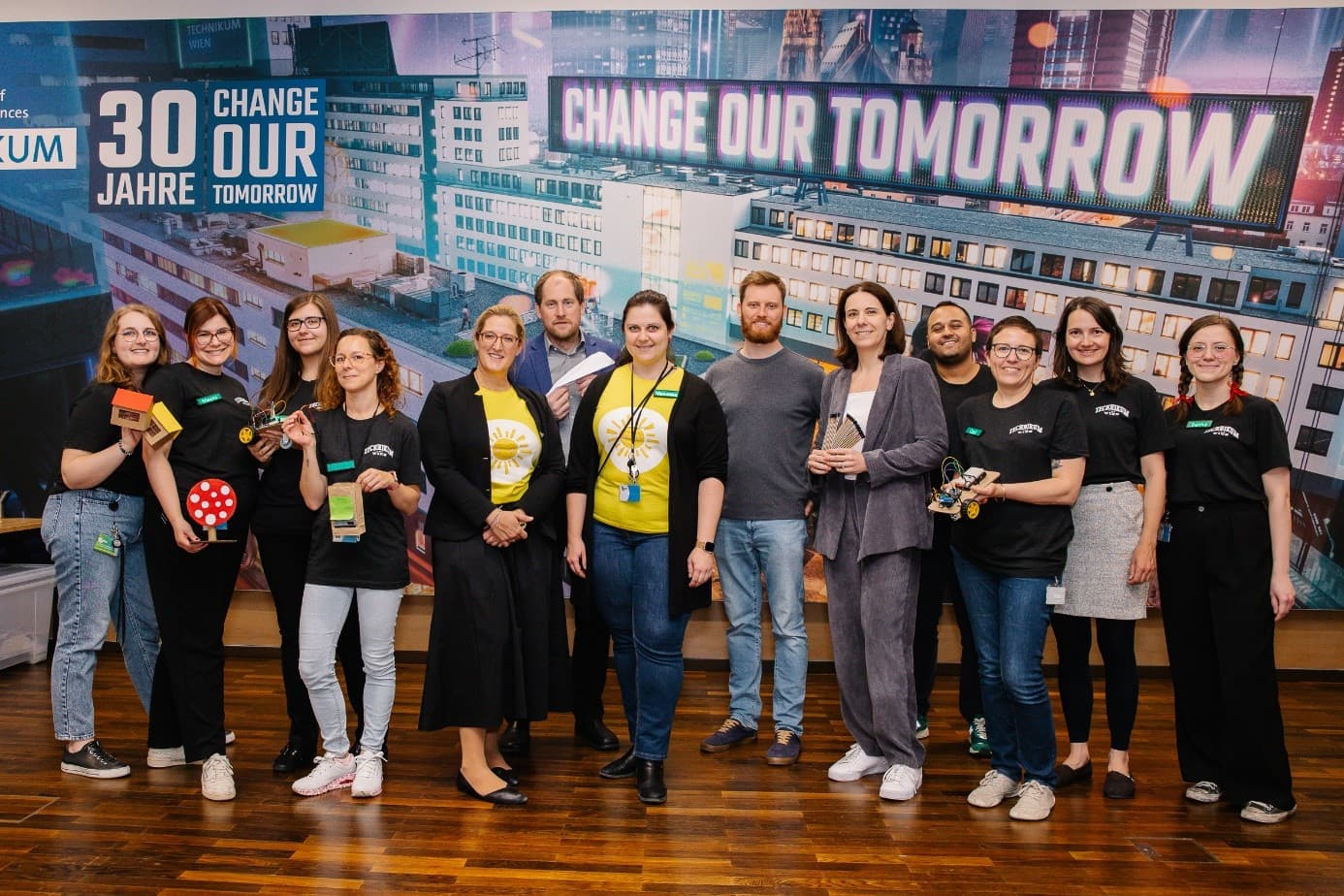 Eine Gruppe von 14 Personen, von denen einige kleine Trophäen oder Requisiten in der Hand halten, steht in einem Innenraum vor einem Transparent mit der Aufschrift '30 Jahre Change Our Tomorrow' zusammen. Die meisten lächeln und sind leger gekleidet.
